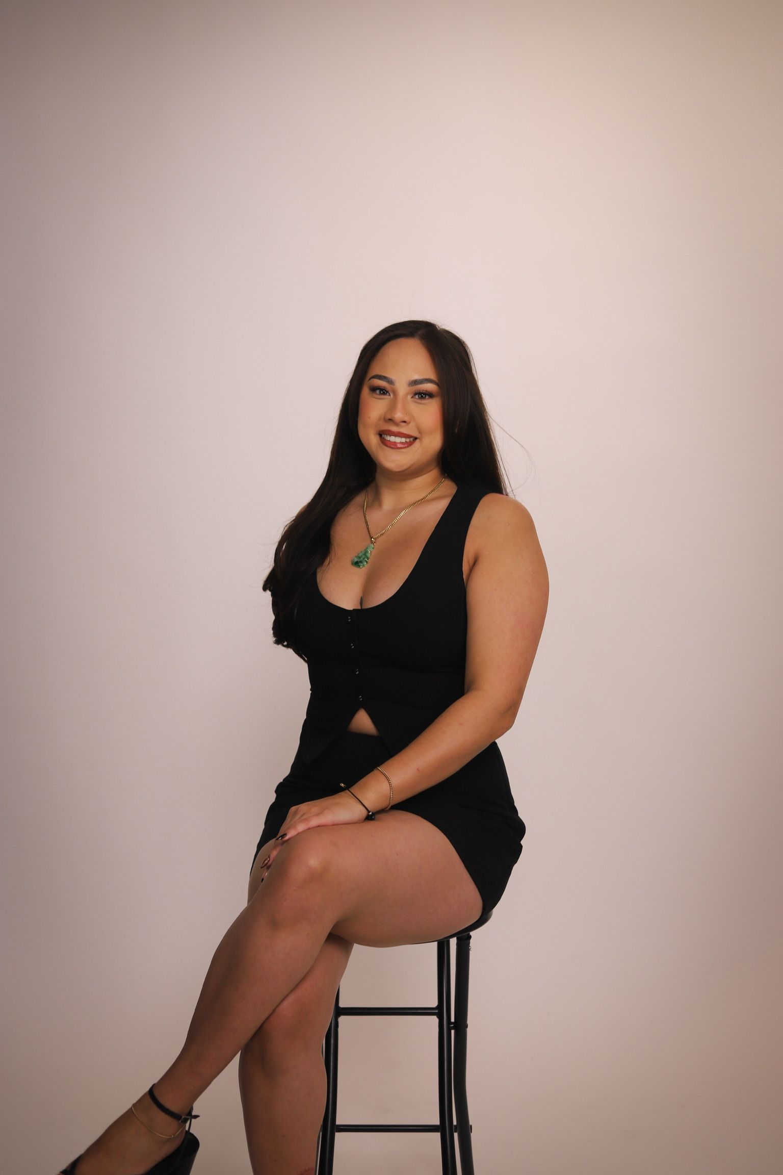 Woman in a black dress sitting on a stool, smiling. She is against a white background.