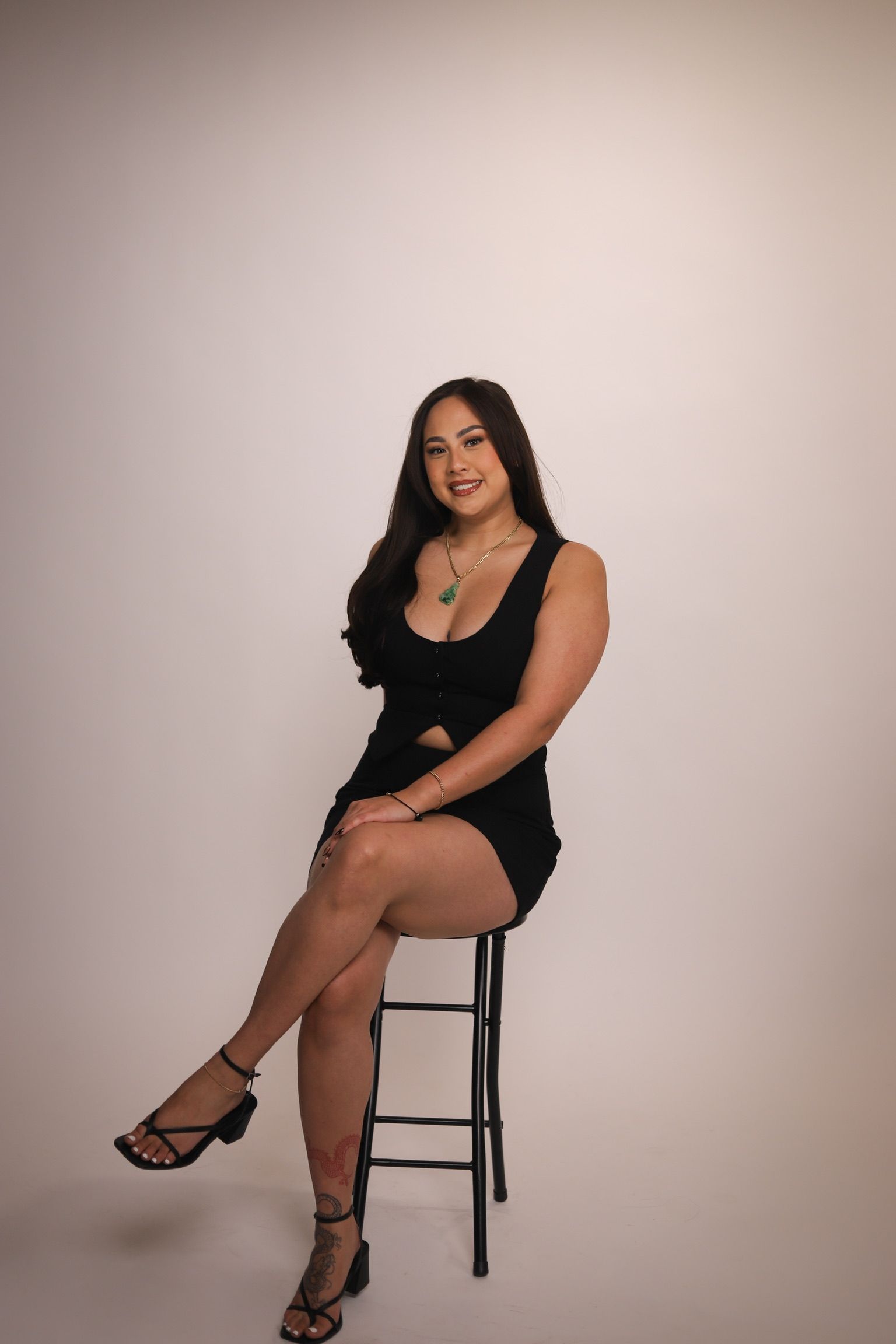 Woman in a black dress sits on a stool, smiling. She wears sandals and a necklace against a white backdrop.