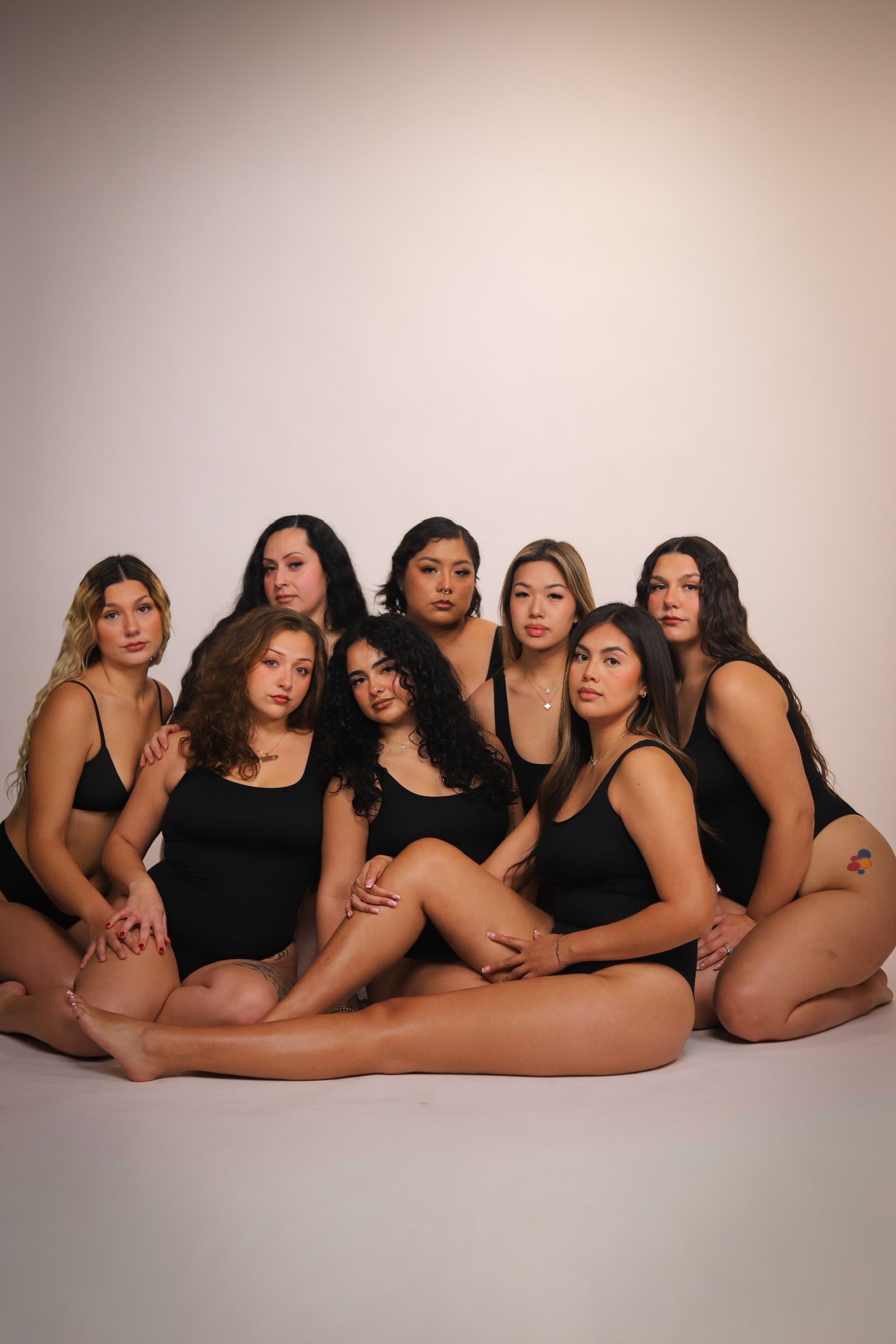Group of diverse women in black bodysuits, posed on the floor against a plain background.