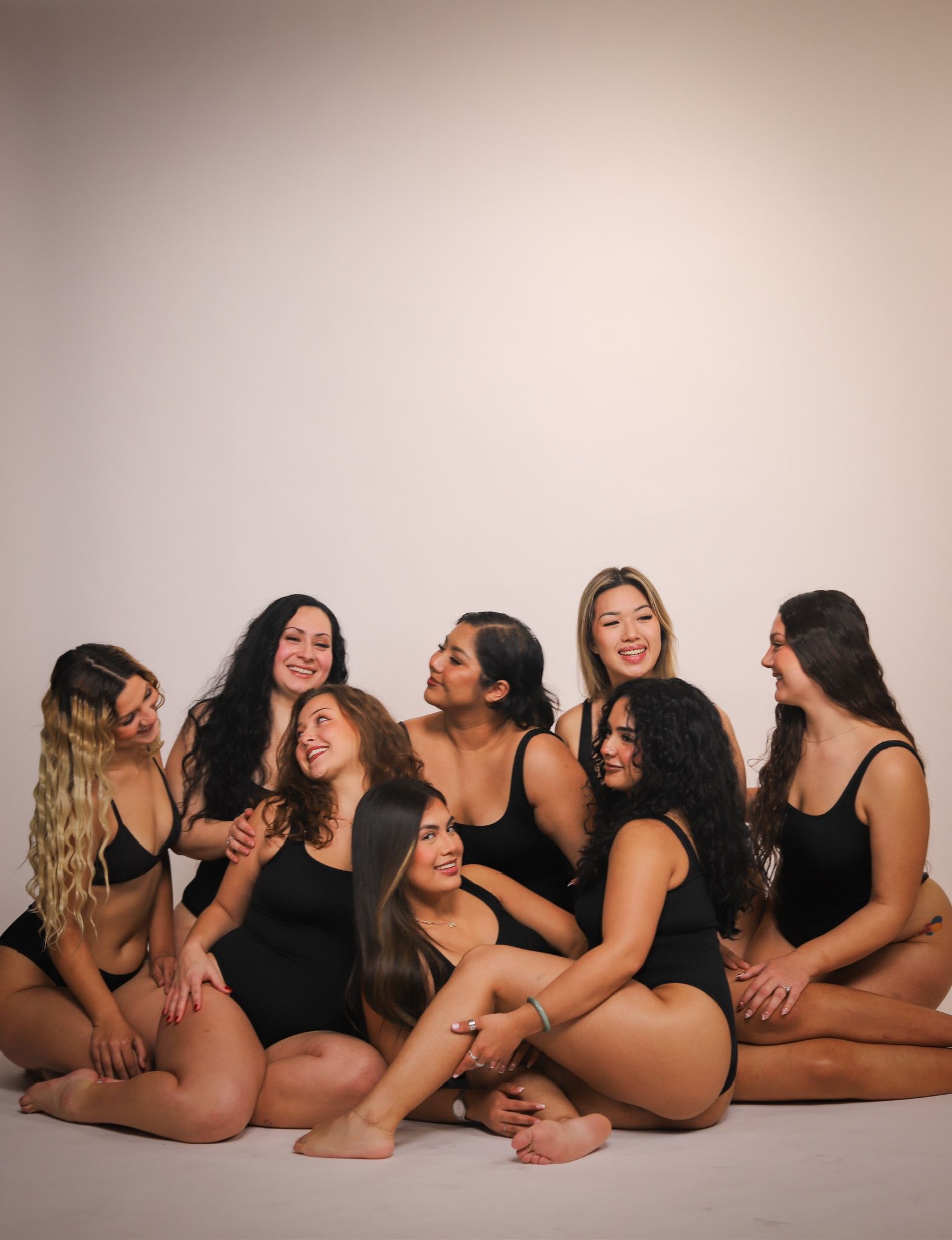 A group of diverse women in black bodysuits, smiling, sitting close together against a white backdrop.