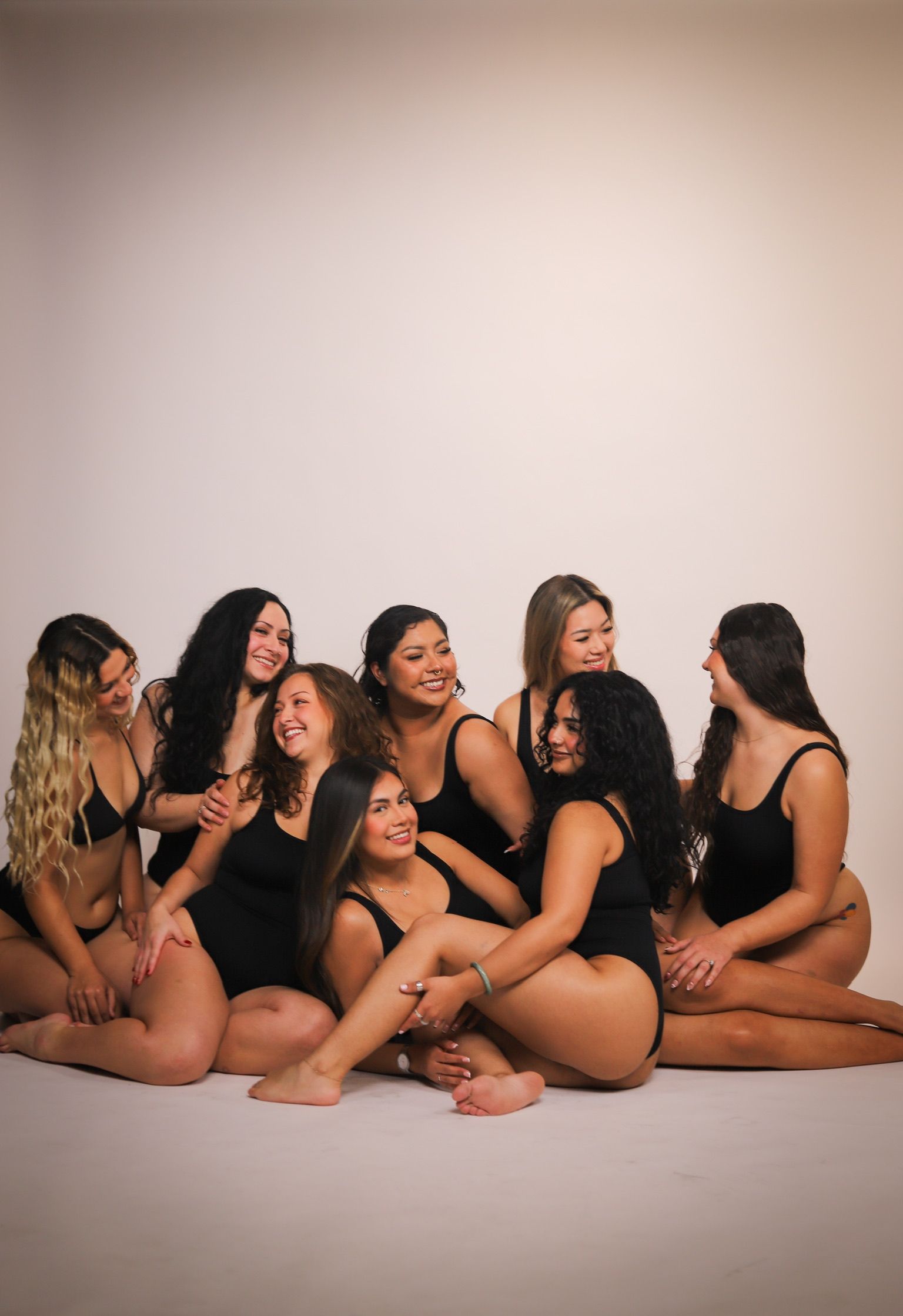 Group of women in black swimwear pose together in studio, smiling and laughing.
