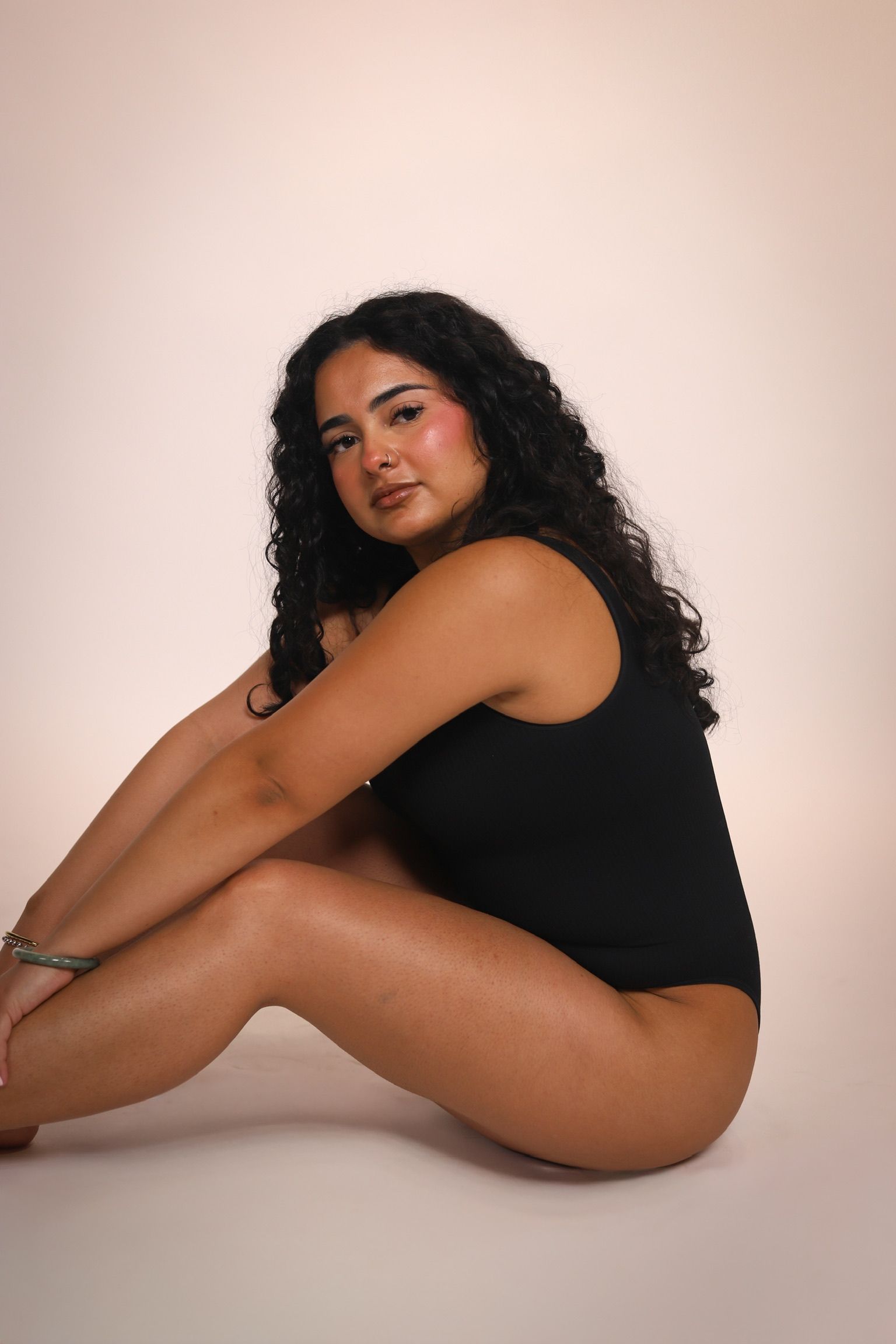 Woman in beige underwear smiles, hands behind head, against a beige background.