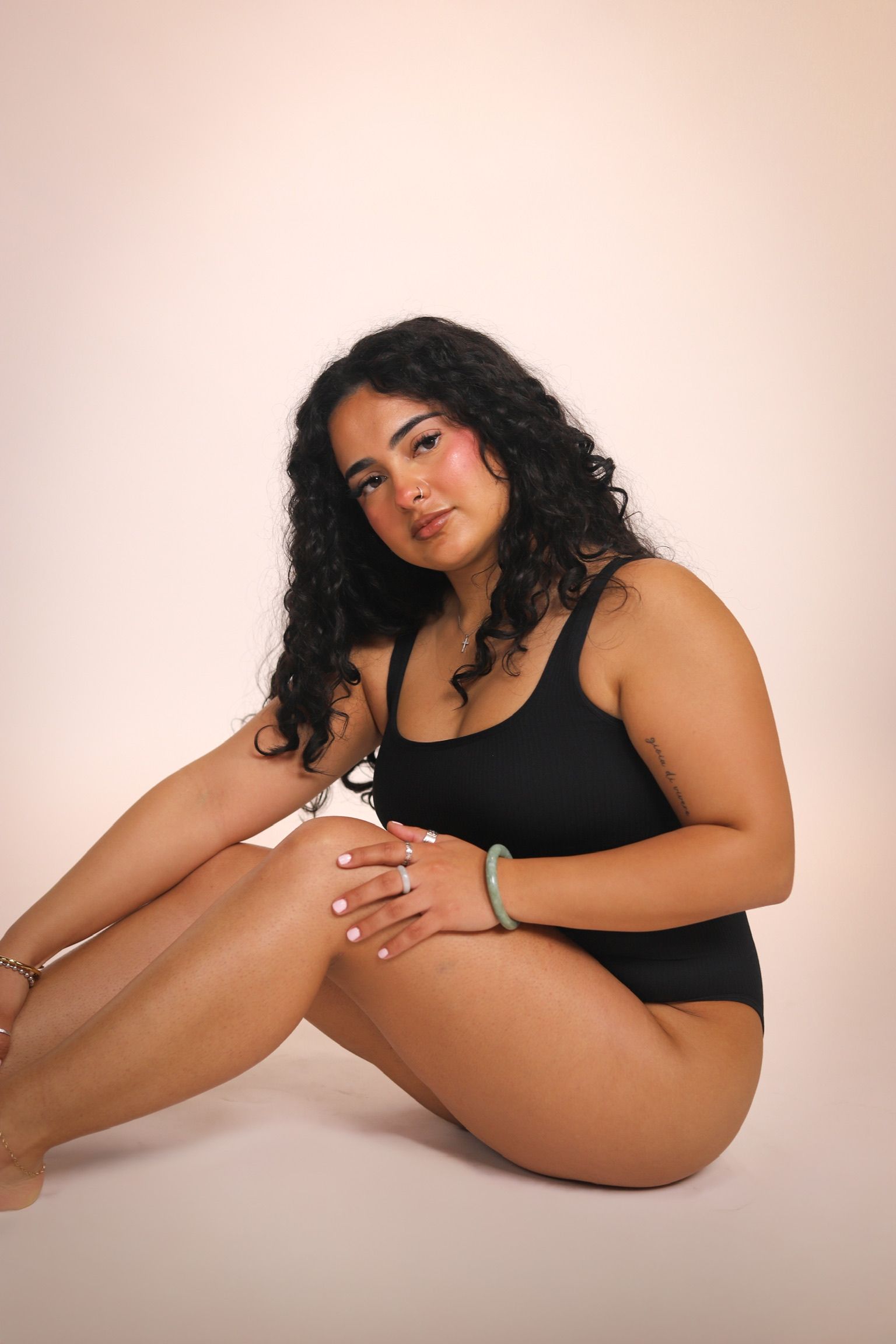 Woman in beige lingerie poses with hands behind head, smiling. Beige background.