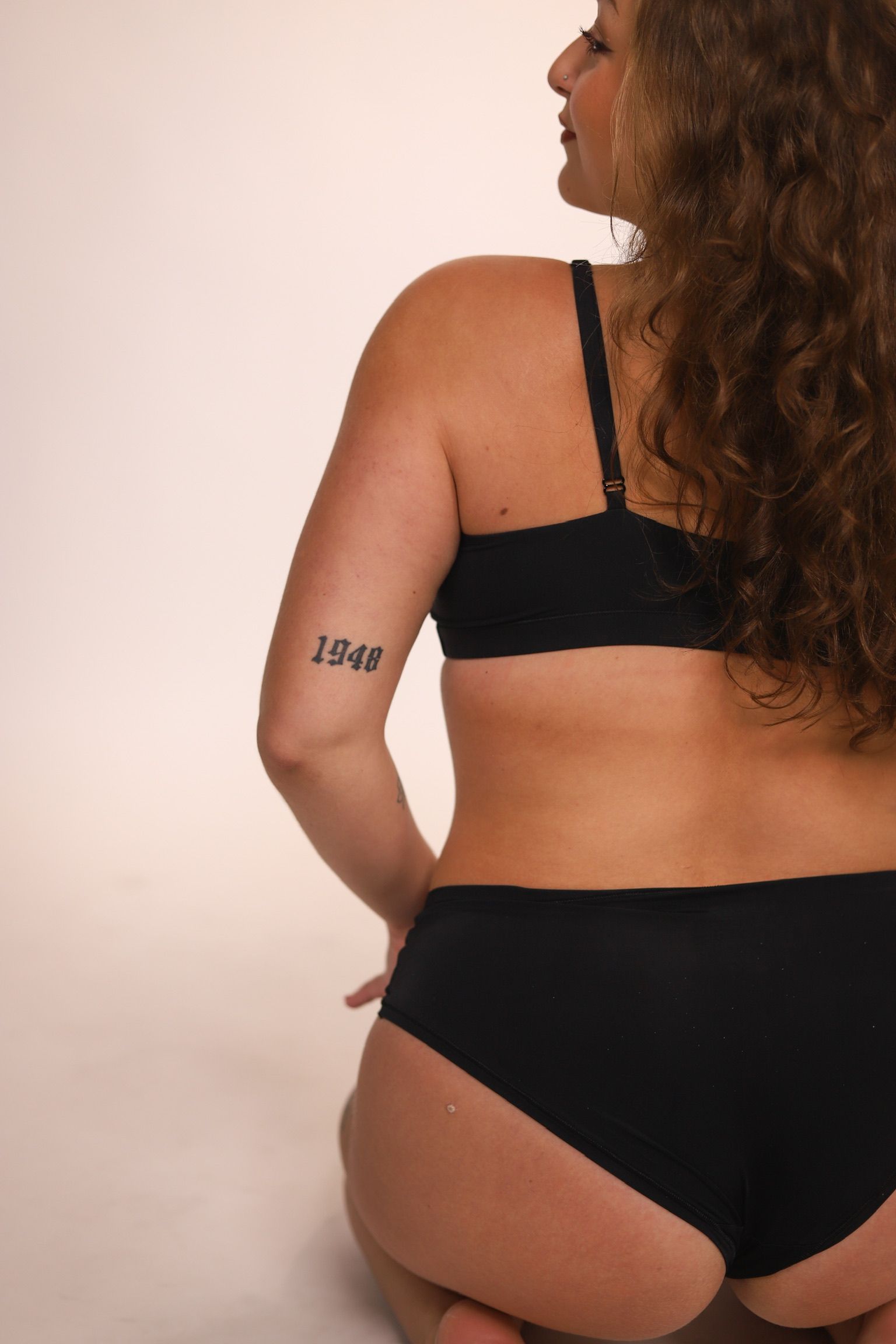 Woman in white top, arm raised, hand touching armpit, smiling, beige background.