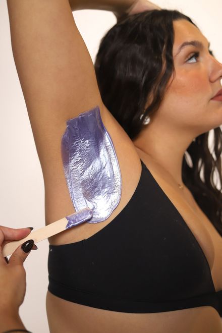 Woman raising arm, hand touching armpit, wearing a white tank top, against a beige background.