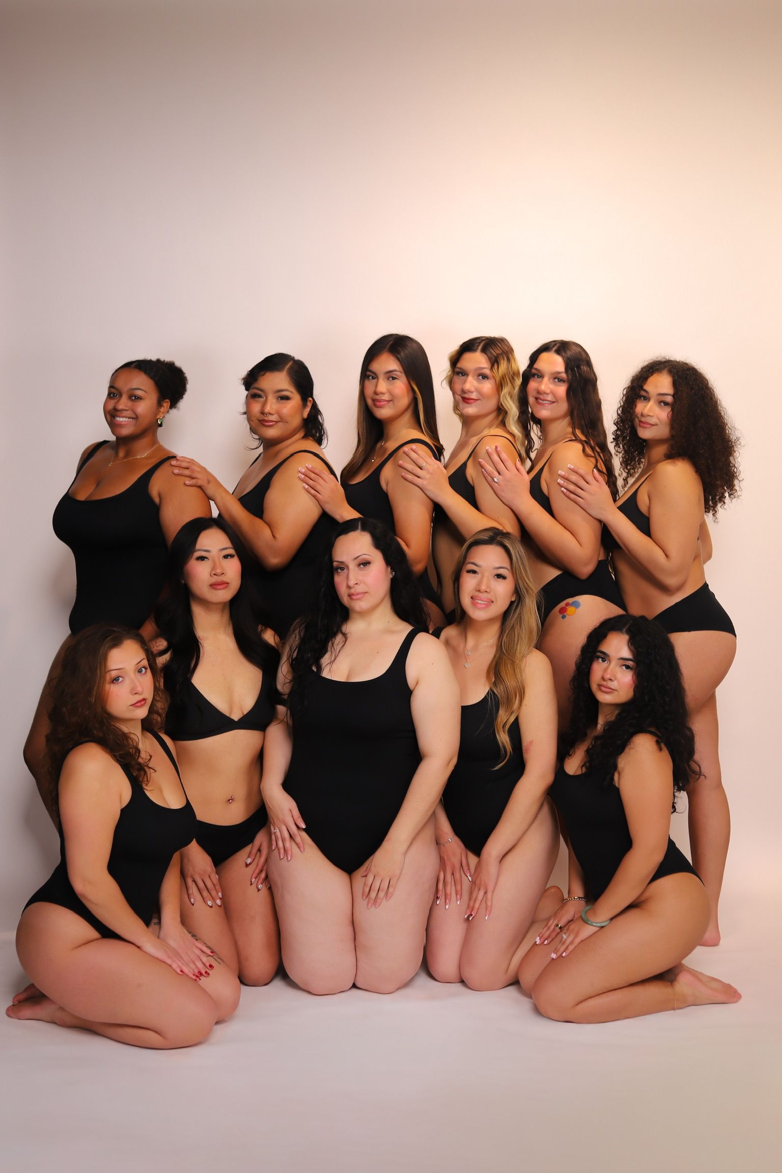 Eleven women in black swimsuits posing against a white background.