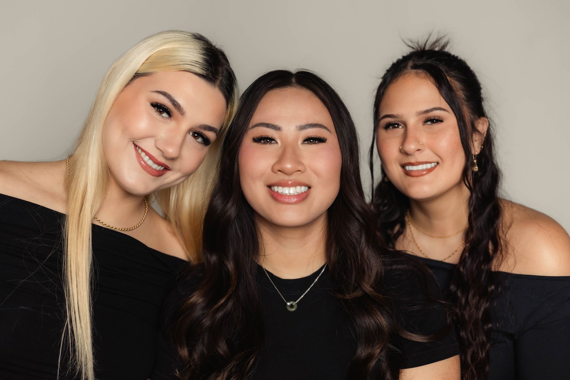 Three smiling women in black tops pose closely together against a light gray background.