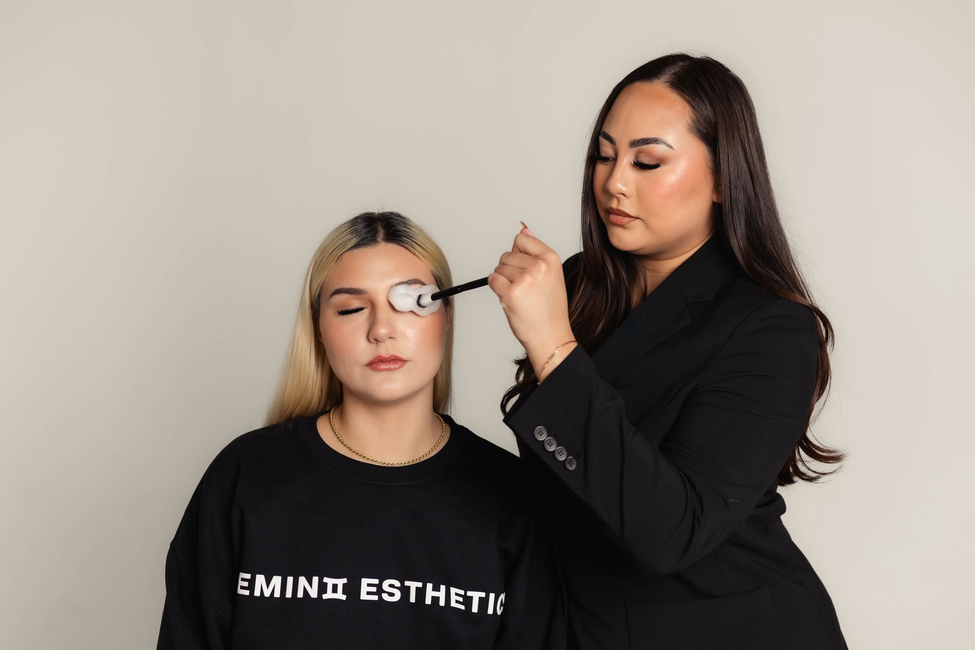 Woman with eye tool examining another woman's eye; both have closed eyes, plain background.