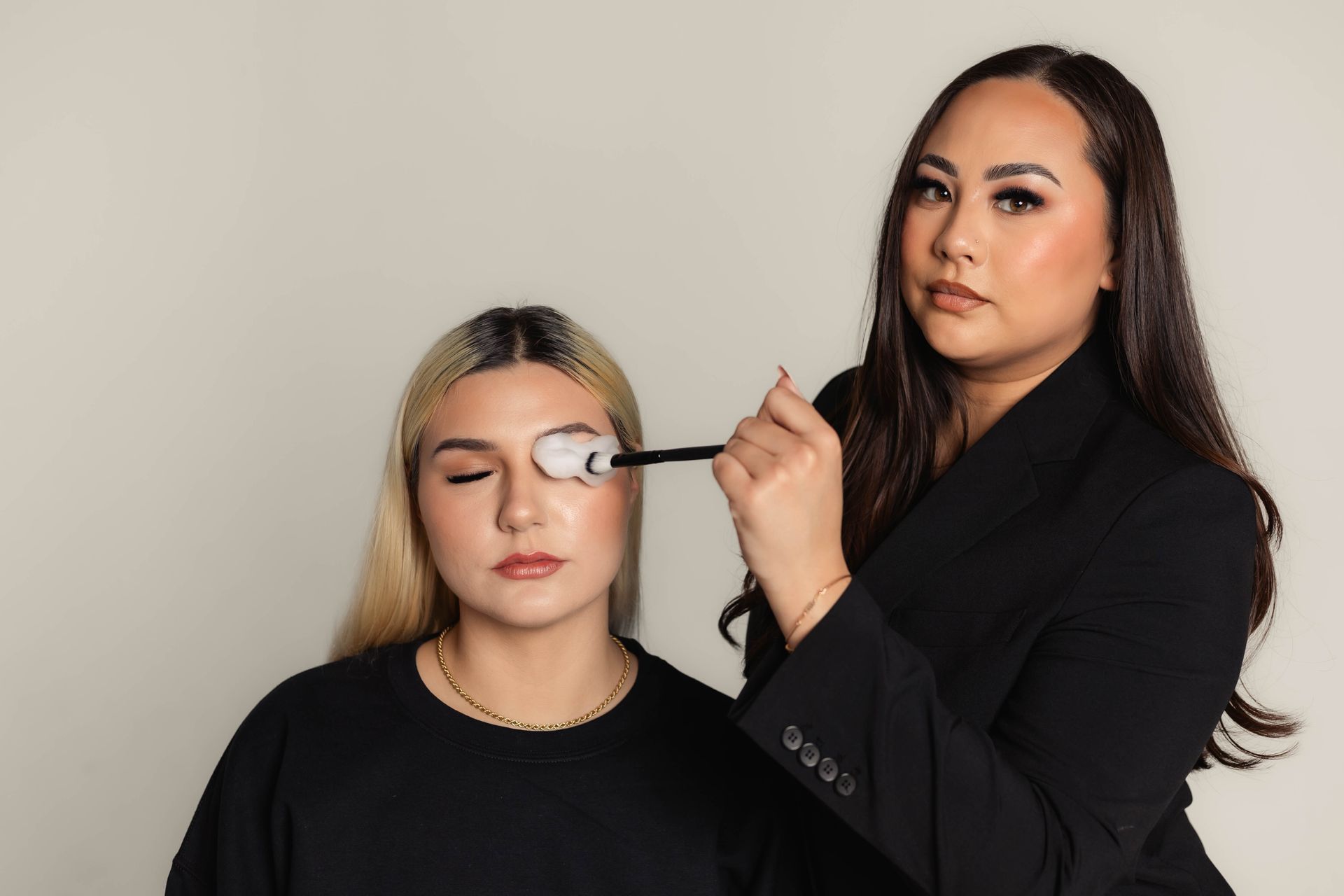 Makeup artist applying eyeshadow to a client's closed eye. Both are in black shirts, against a light backdrop.