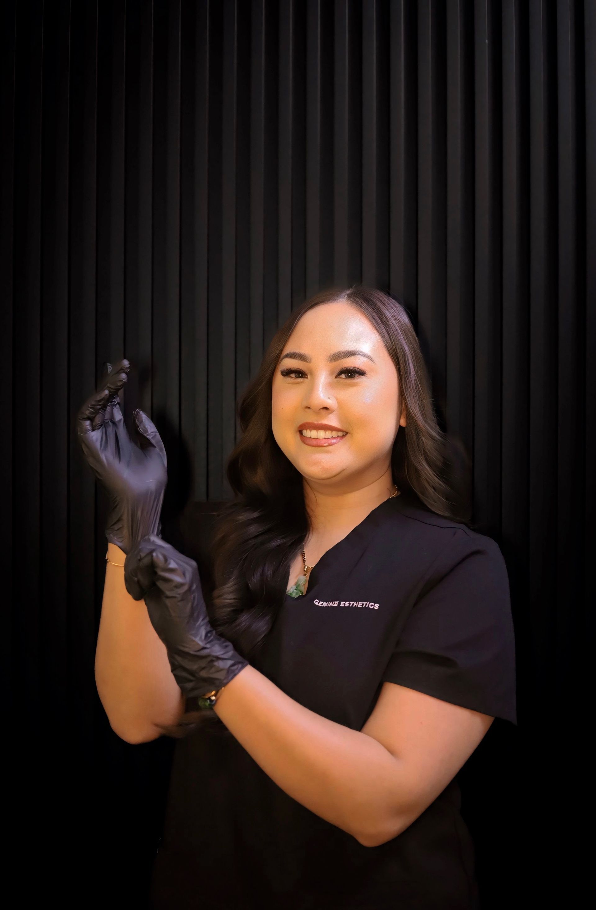 Woman in black scrubs and gloves smiles, stands against black textured wall.