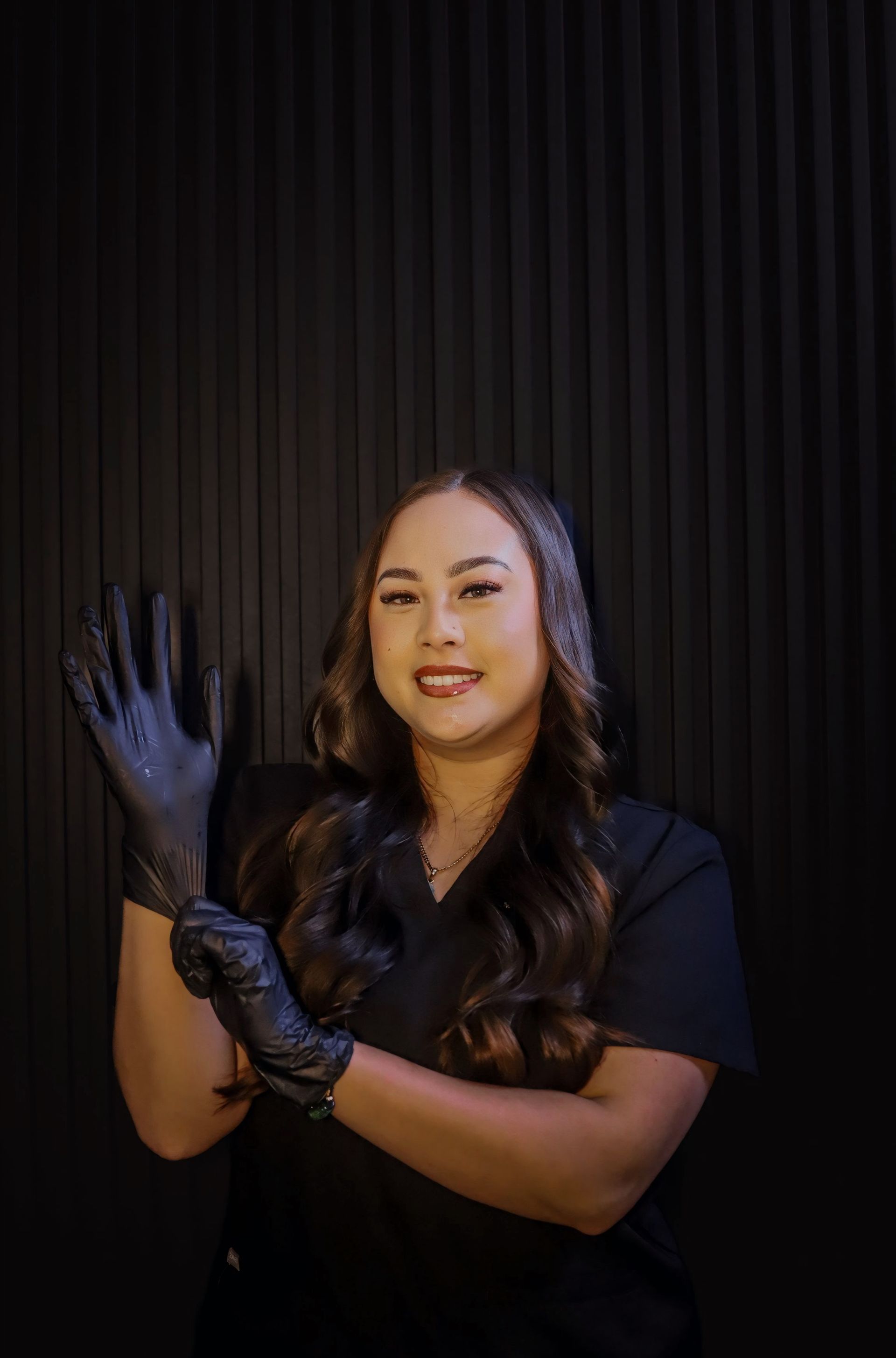 Woman in black scrubs putting on a black glove, smiling at the camera against a textured black background.