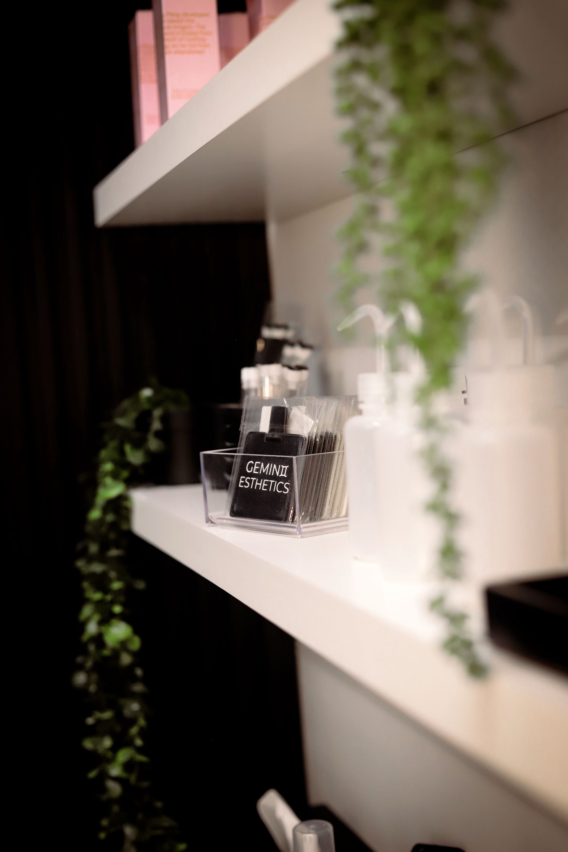 White shelves holding cosmetic products, clear containers, and hanging greenery in a spa setting.