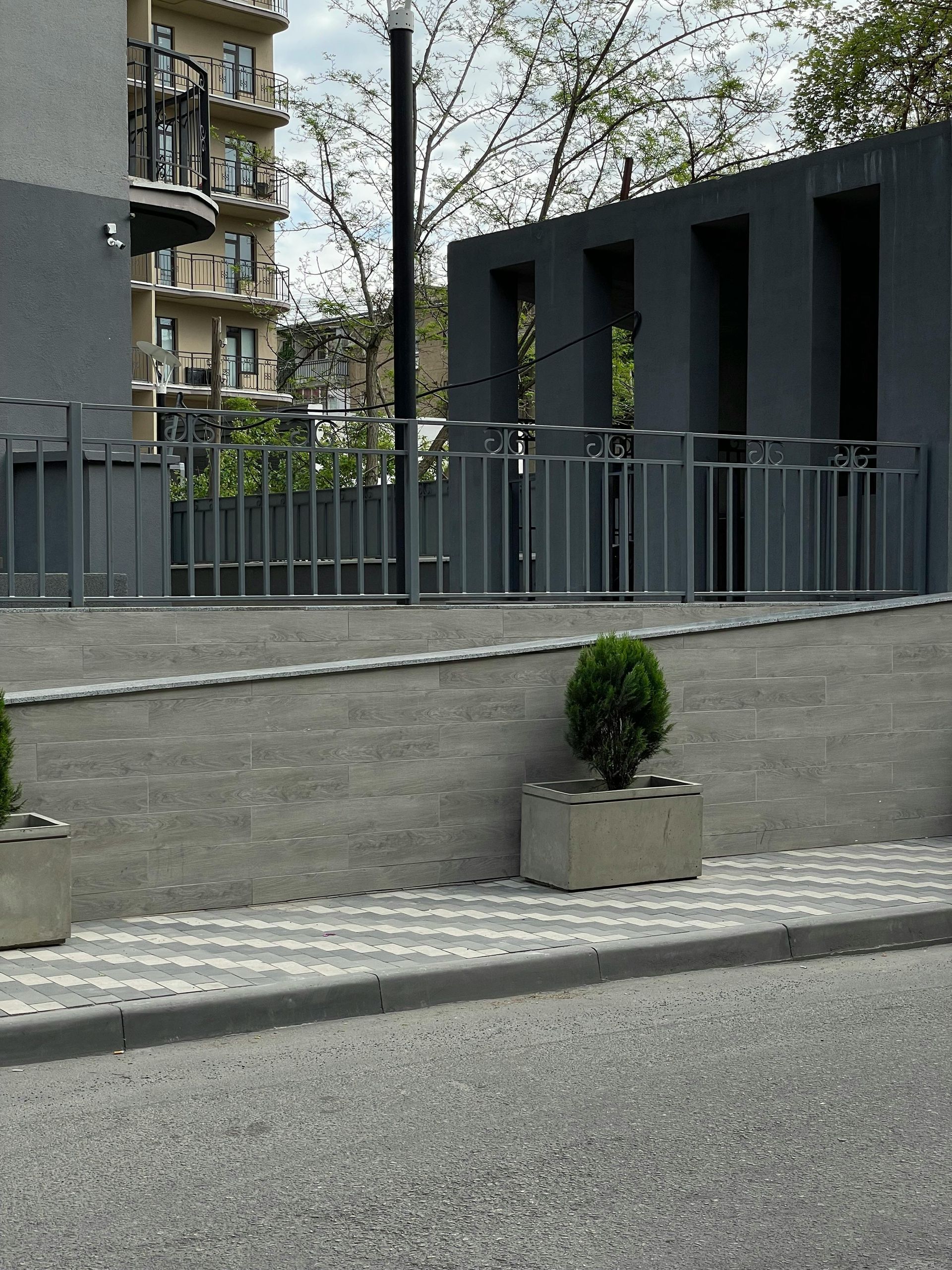 Gray concrete building exterior with a decorative fence, planters, and a patterned sidewalk.