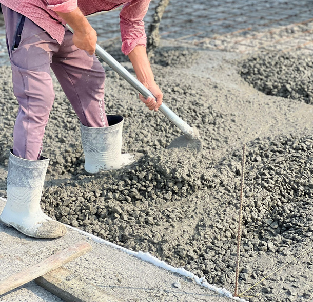 Person in pink shirt and pants, white boots, shoveling concrete mix.