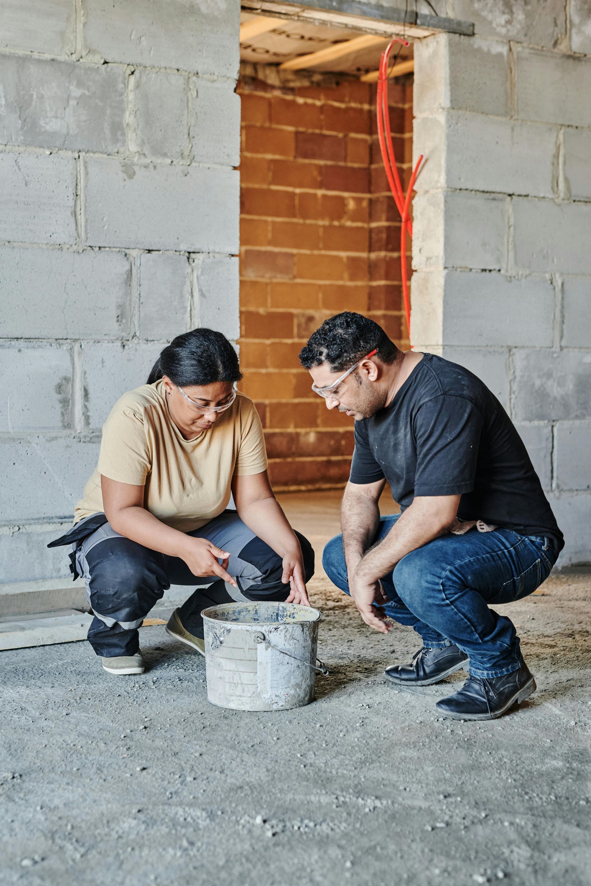 Two people in work clothes crouch near a bucket, inspecting its contents in a construction site doorway.