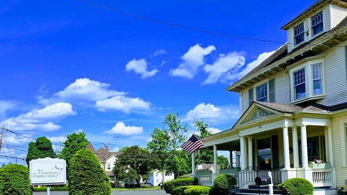 A two-story house with a porch under a bright blue sky. A sign and American flag are in front.