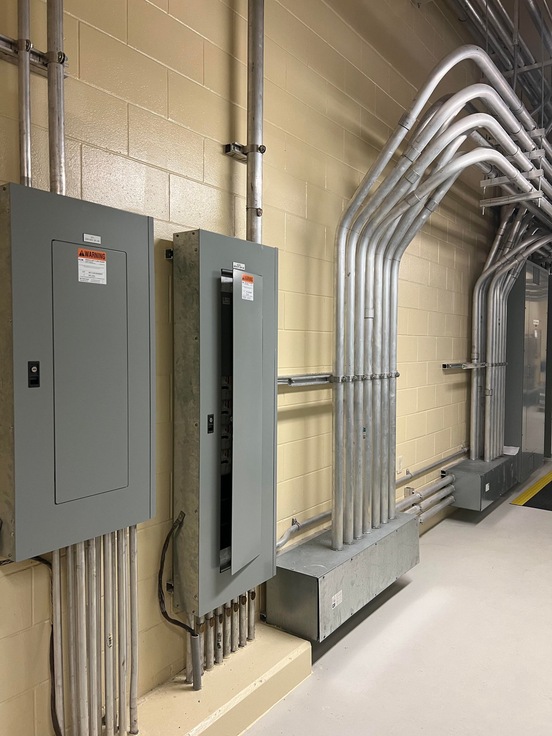 Smiling electrician in blue coveralls and hard hat, holding a multimeter near an electrical panel.