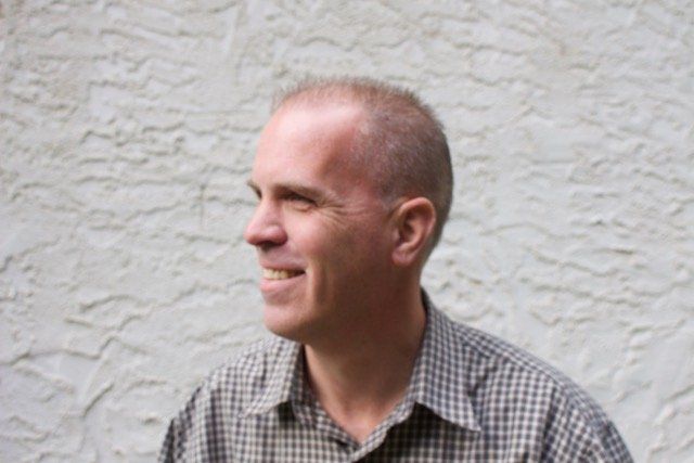 A man in a plaid shirt is smiling in front of a white wall