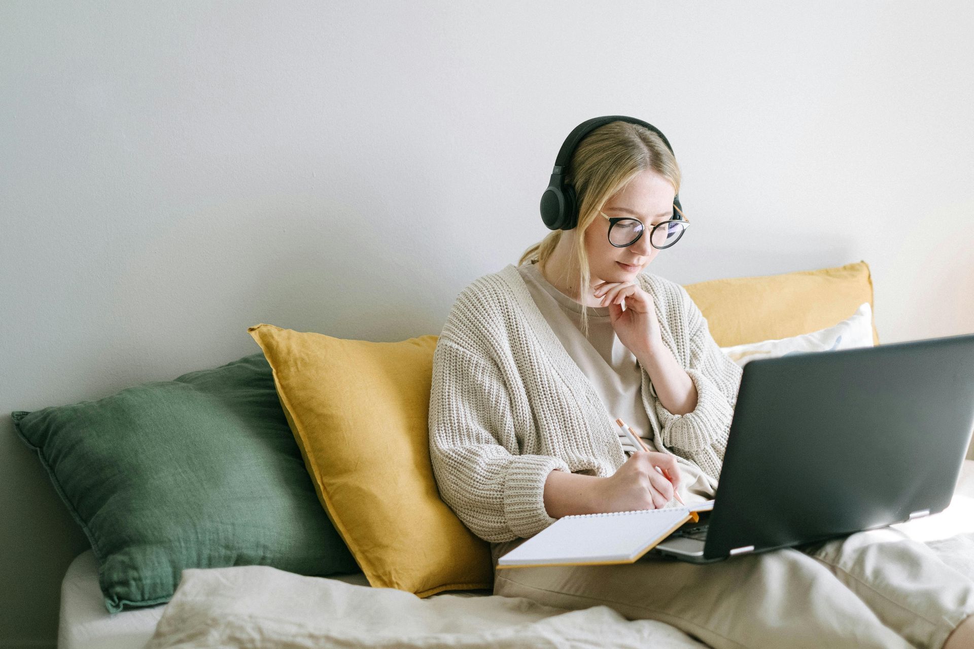 Woman with glasses and headphones, writing in a notebook while looking at a laptop on a bed.