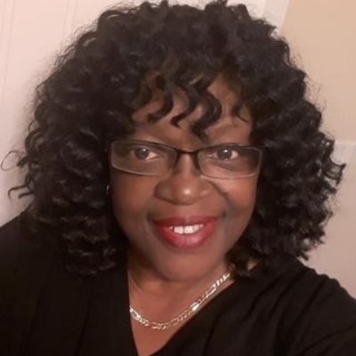 Woman with black curly hair, glasses, red lipstick, smiling. Wearing black shirt, silver necklace.