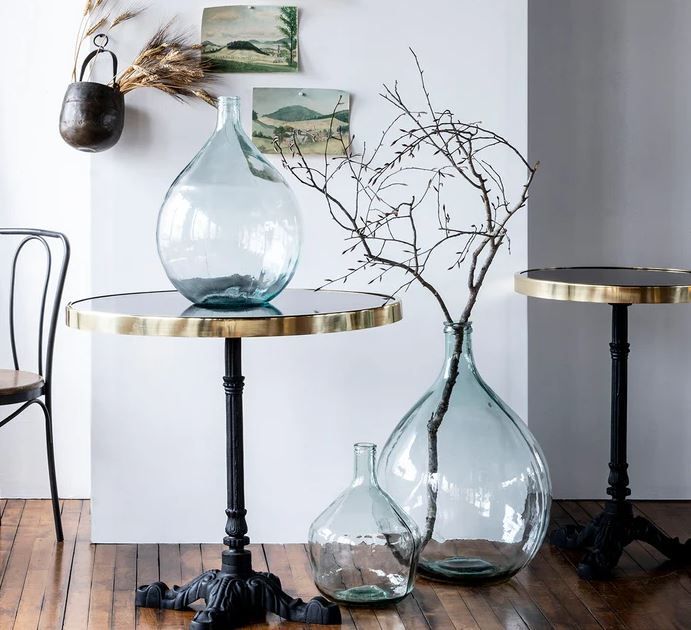 Three glass vases on round brass-rimmed tables. One vase holds branches. White wall background.
