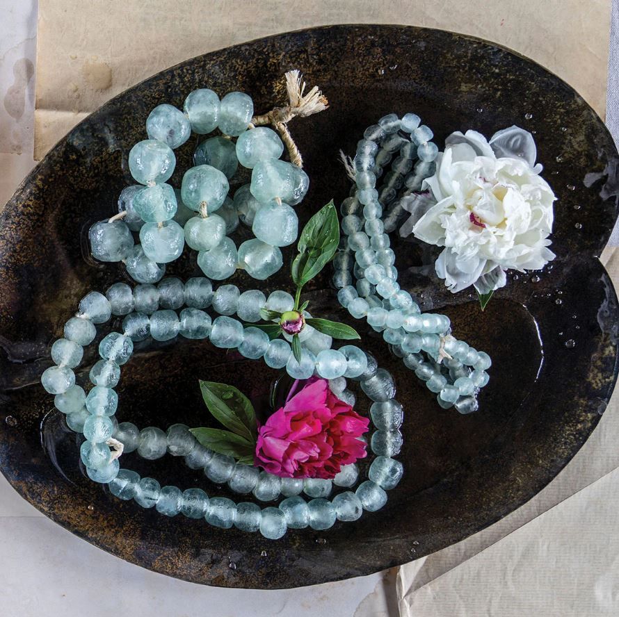 Beaded necklaces and flowers in a rustic, dark bowl. Soft blue beads with pink and white flowers.