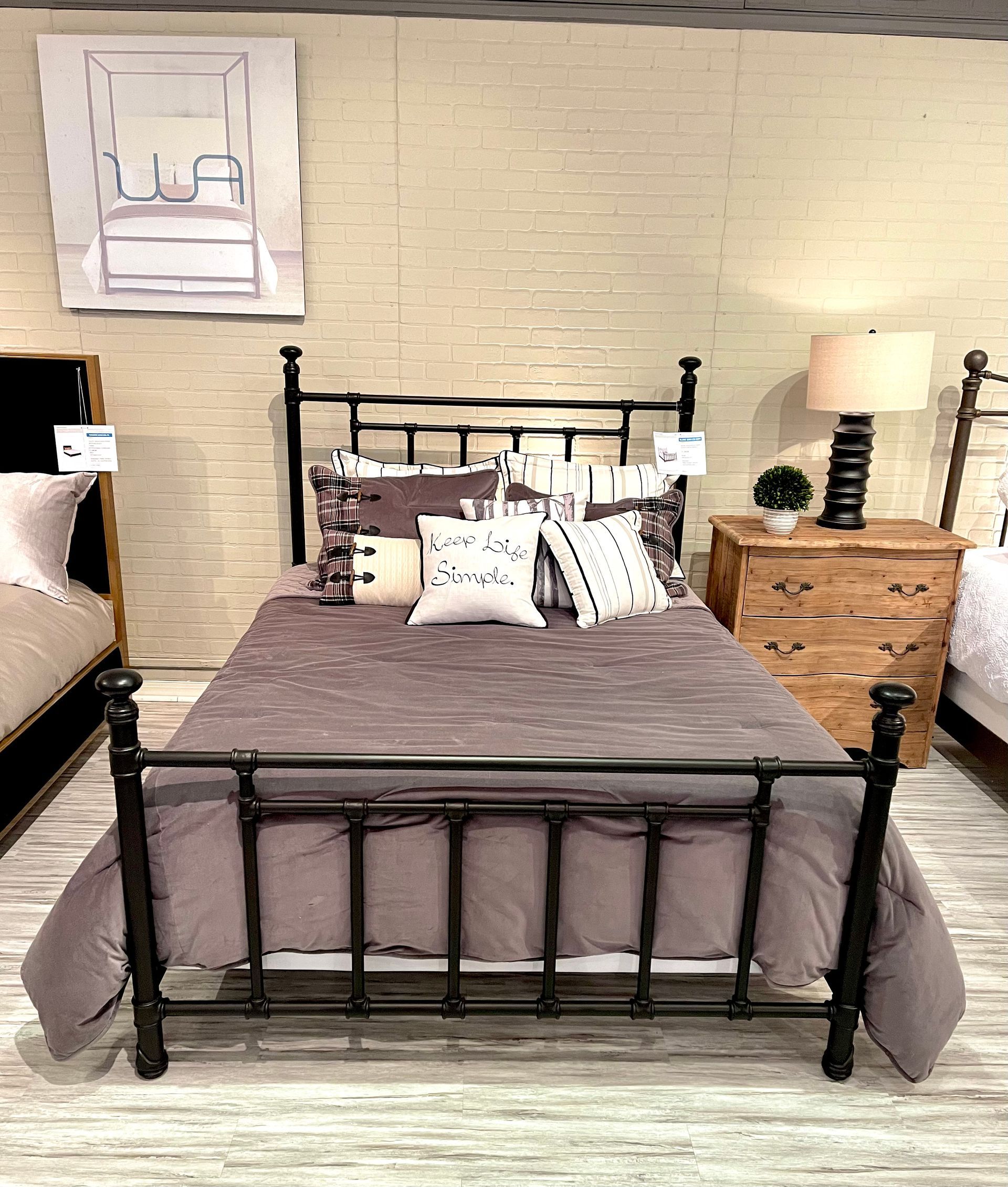 Metal bed frame with a gray comforter and pillows against a light brick wall.