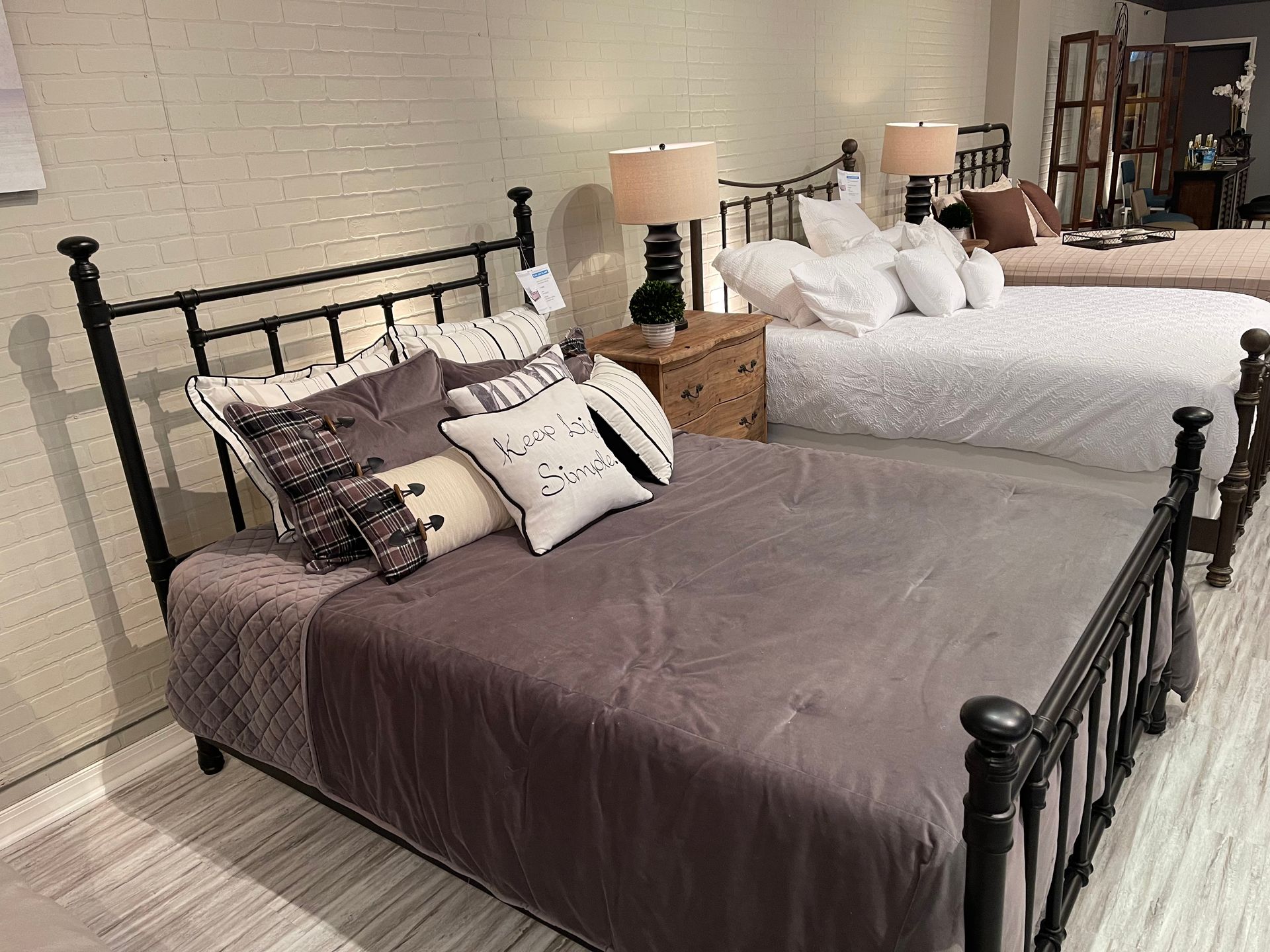 Three black metal beds with bedding in a showroom setting; white brick wall backdrop.