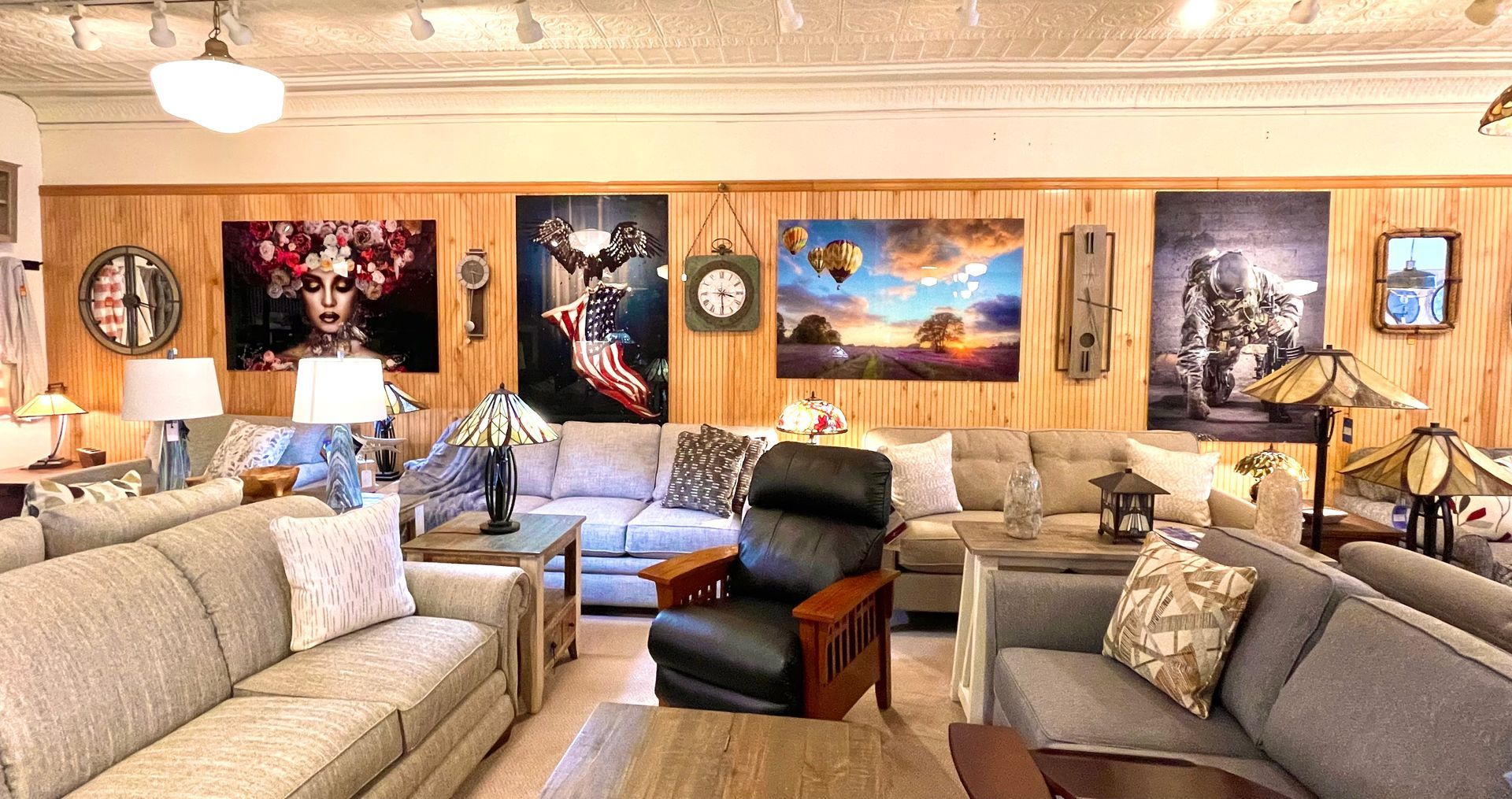 Interior of a furniture store with sofas, a recliner, tables, lamps, and wall art in a warm-toned room.