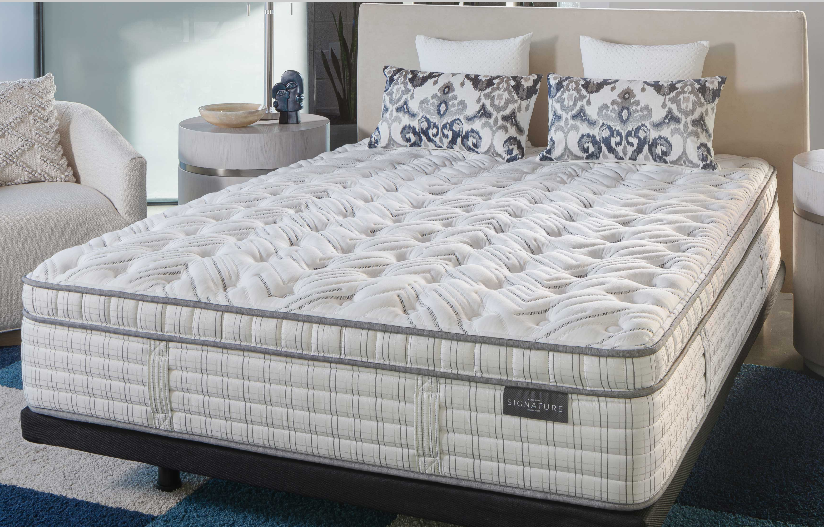 Queen-sized bed with quilted mattress, patterned pillows, and gray trim, in a bedroom setting.