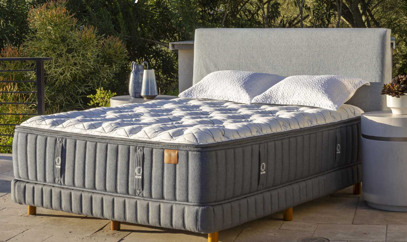 Gray bed with white bedding, on a patio. Includes a headboard, pillows, and a side table.