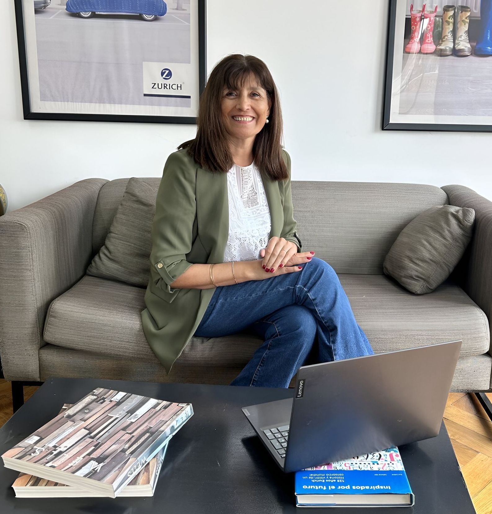 Mujer sentada en un sofá, vestida con un blazer verde y jeans, sonriendo, con una computadora portátil y libros sobre una mesa.