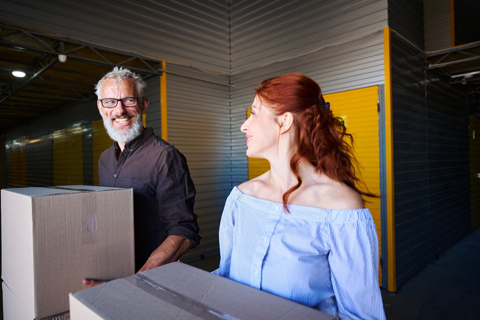 A man and woman carrying boxes in a storage unit. The man smiles, while the woman looks at him.