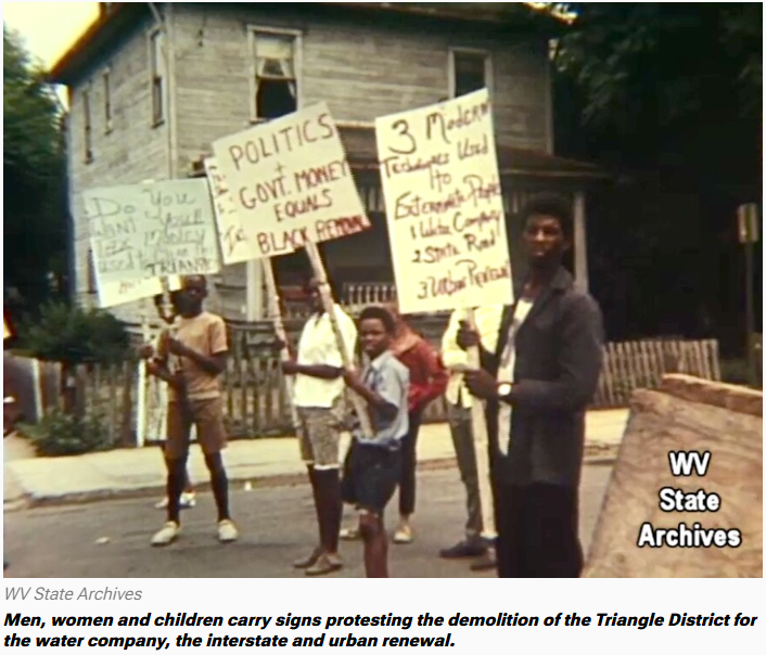 Protesters with signs, including children, in front of a house, opposing demolition for a water company and urban renewal.