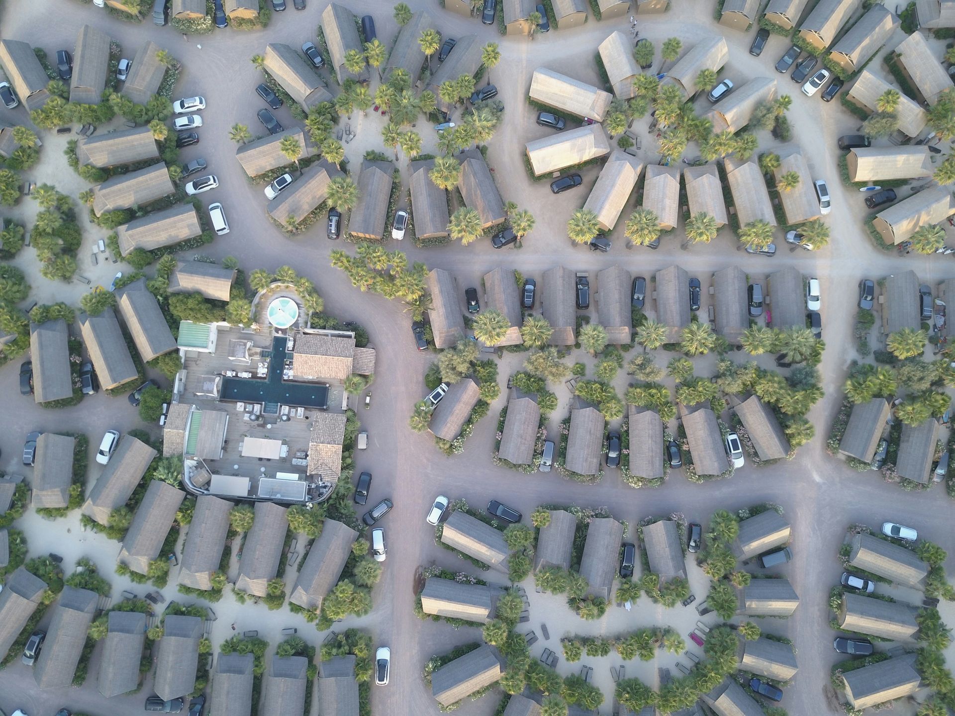 Aerial view of a resort with many bungalows and cars, surrounded by palm trees and a central building.