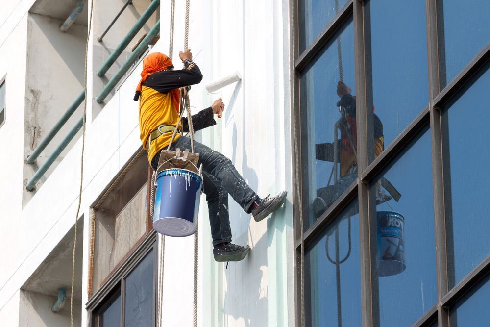 A Man Is Painting A Building With A Roller While Hanging From A Rope — Mid Coast Coatings In Kempsey, NSW