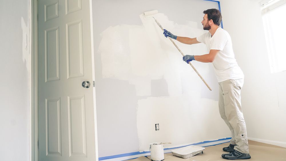 A Man Is Painting A Wall In A Room With A Roller — Mid Coast Coatings In Port Macquarie, NSW