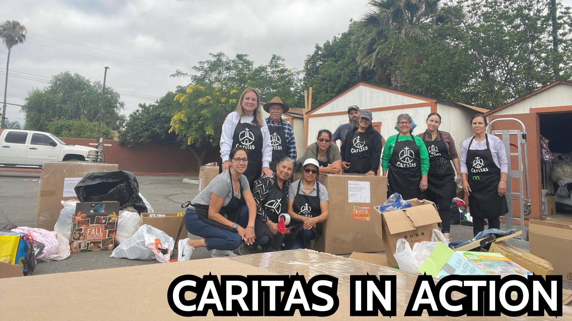 A group of volunteers in black aprons organize boxes and supplies outdoors, with the text 