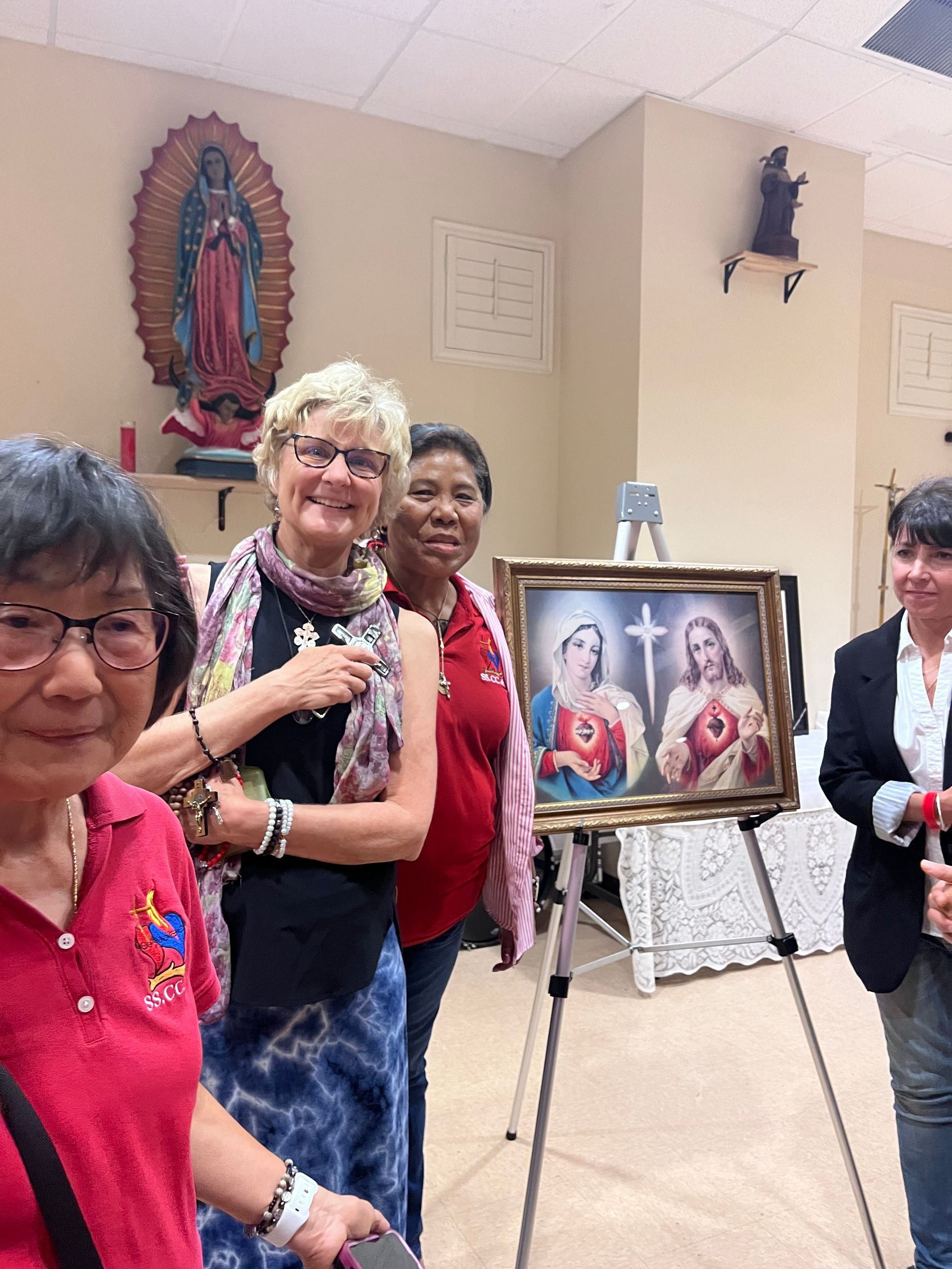 Four people stand in a room, three posing beside a framed painting of the Sacred Heart and Immaculate Heart of Mary.