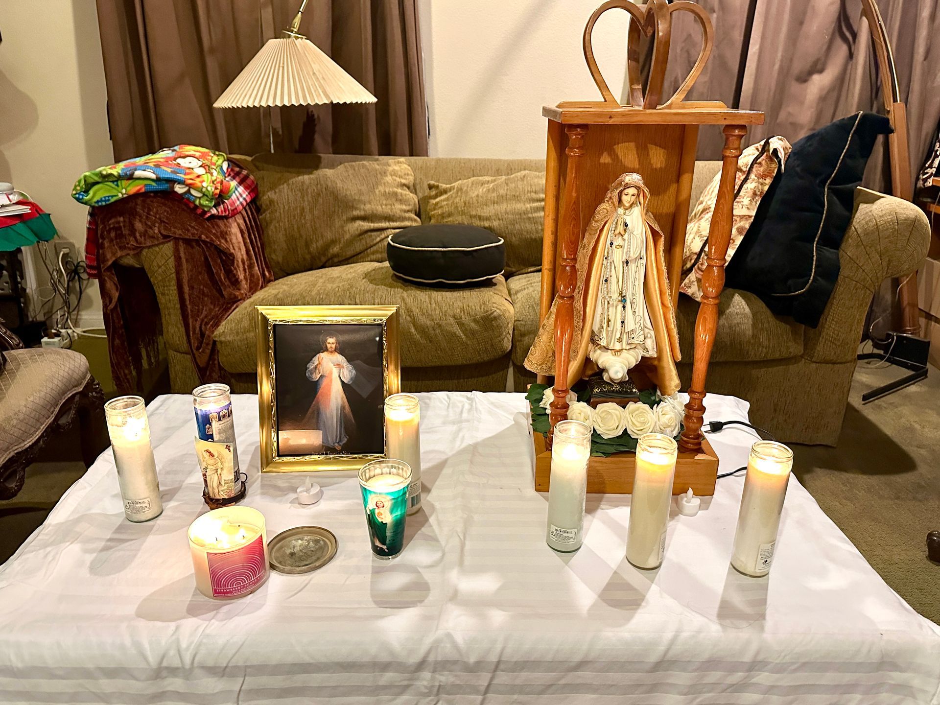 A home altar with a statue of the Virgin Mary, a Divine Mercy framed print, and several lit candles on a white cloth.