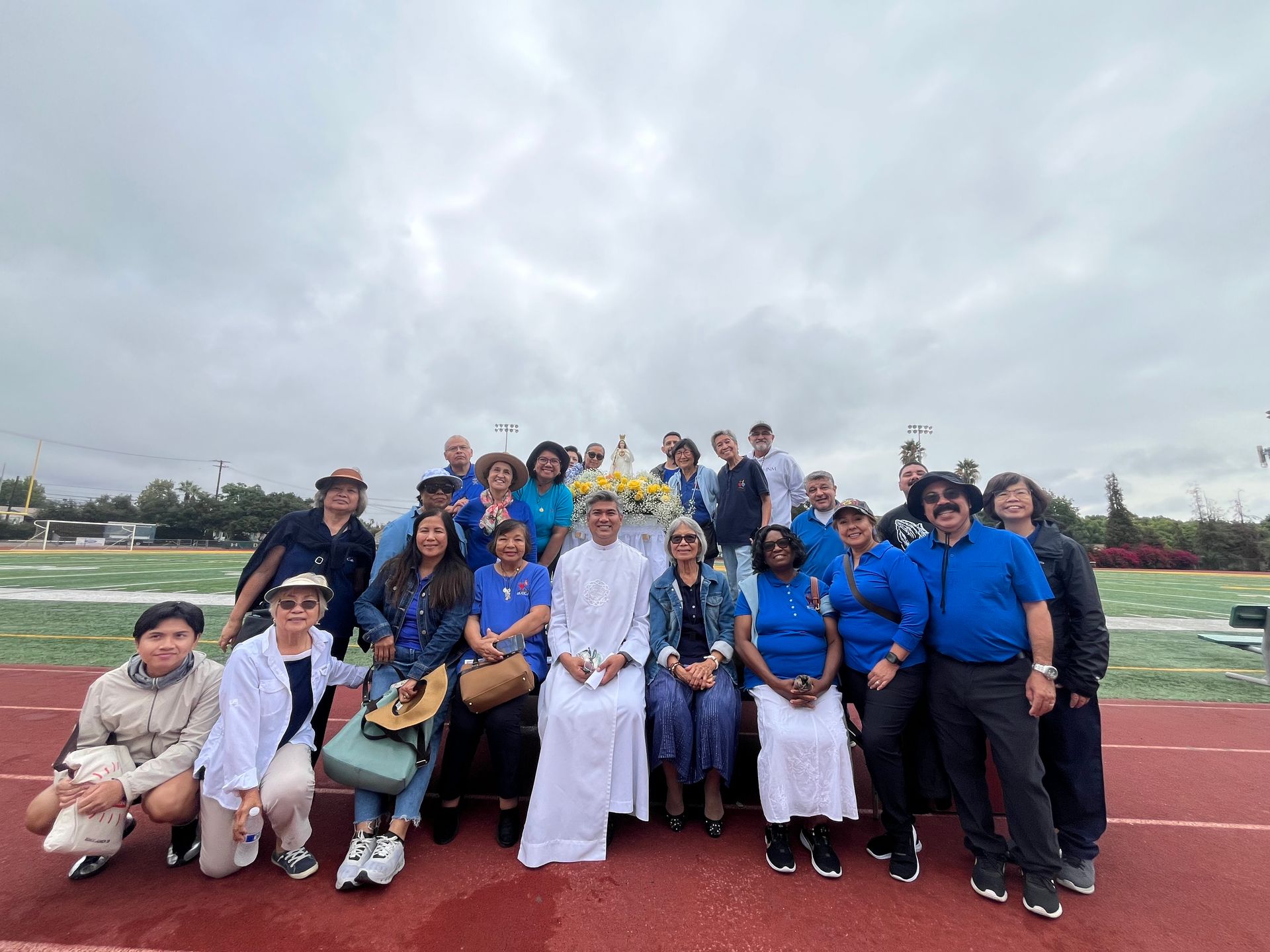 A large group gathers for a photo on a red track with a field in the background. A person in white robes sits in the center.