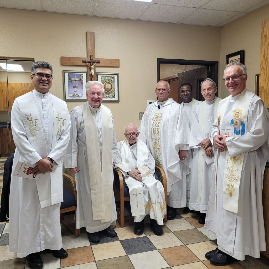 Seven clergy members in liturgical robes stand and sit in a tiled room with a wooden cross on the wall.