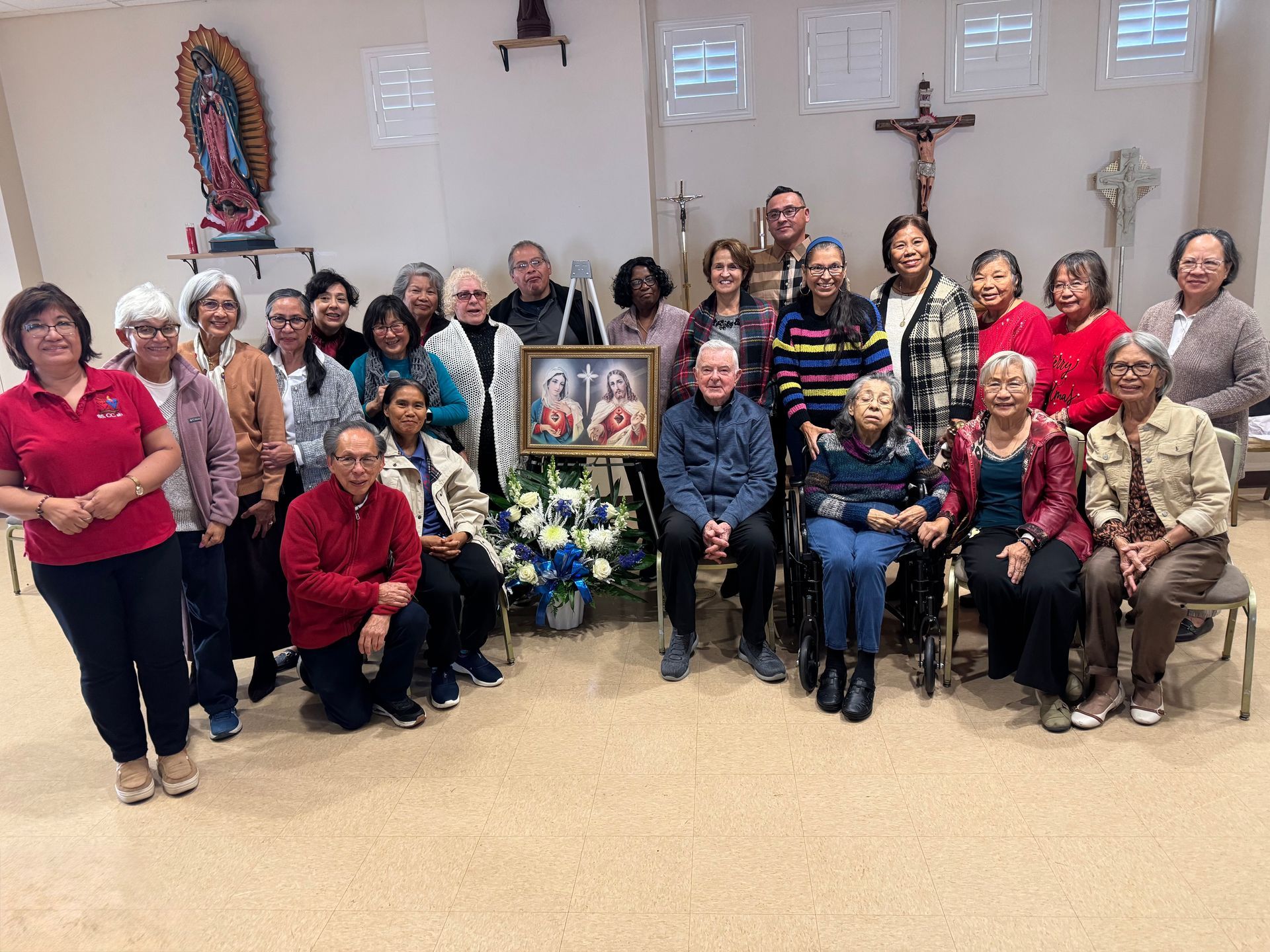 A group of people standing and sitting in a room around a framed religious image, with religious wall decorations.