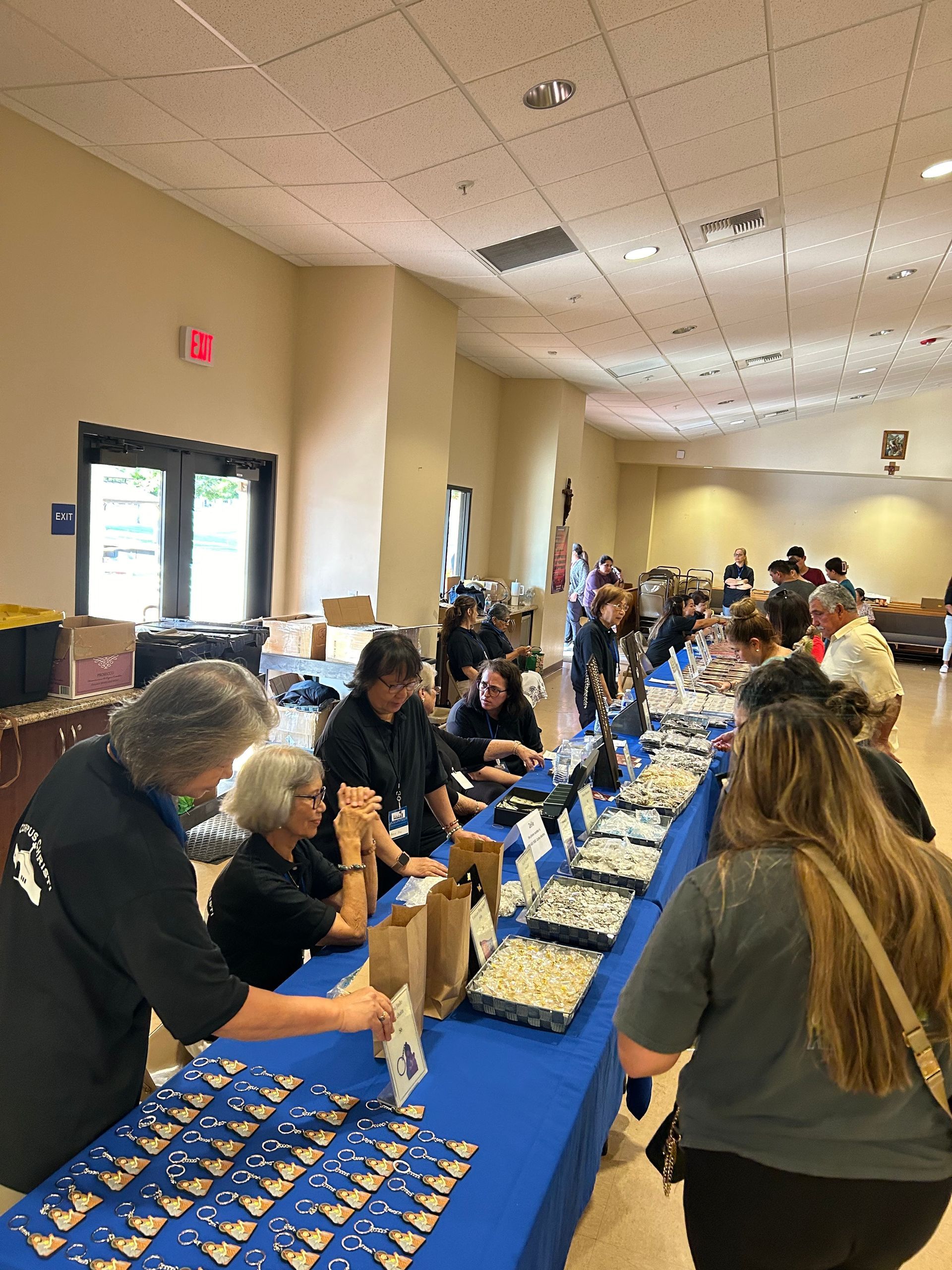 A line of people in a bright room prepare food items along a long table covered in a blue cloth.