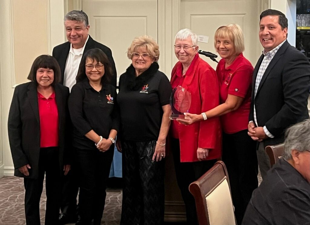 A group of people in business-casual attire stand together in an interior space, with one person holding an award.