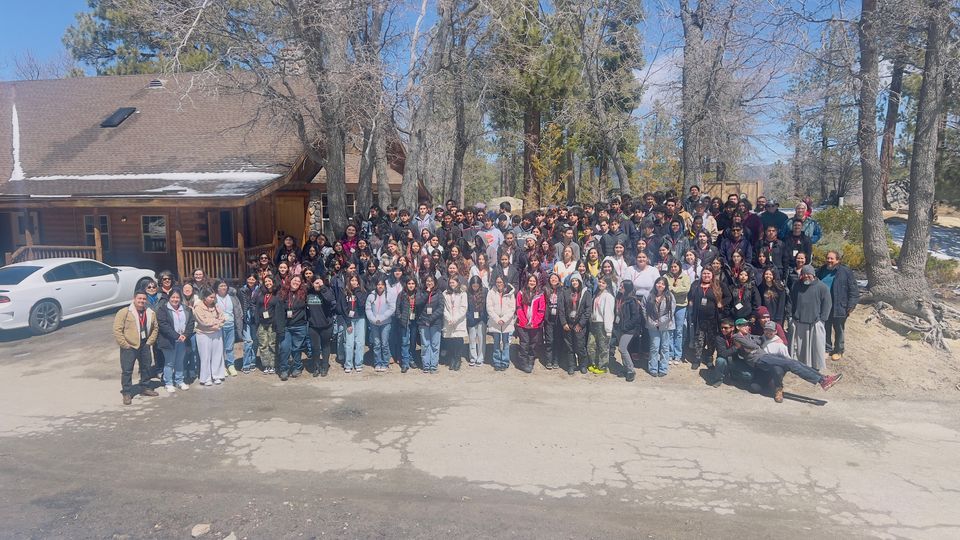 Large group of people posing in front of a wooden building in a wooded area. Sunny day.