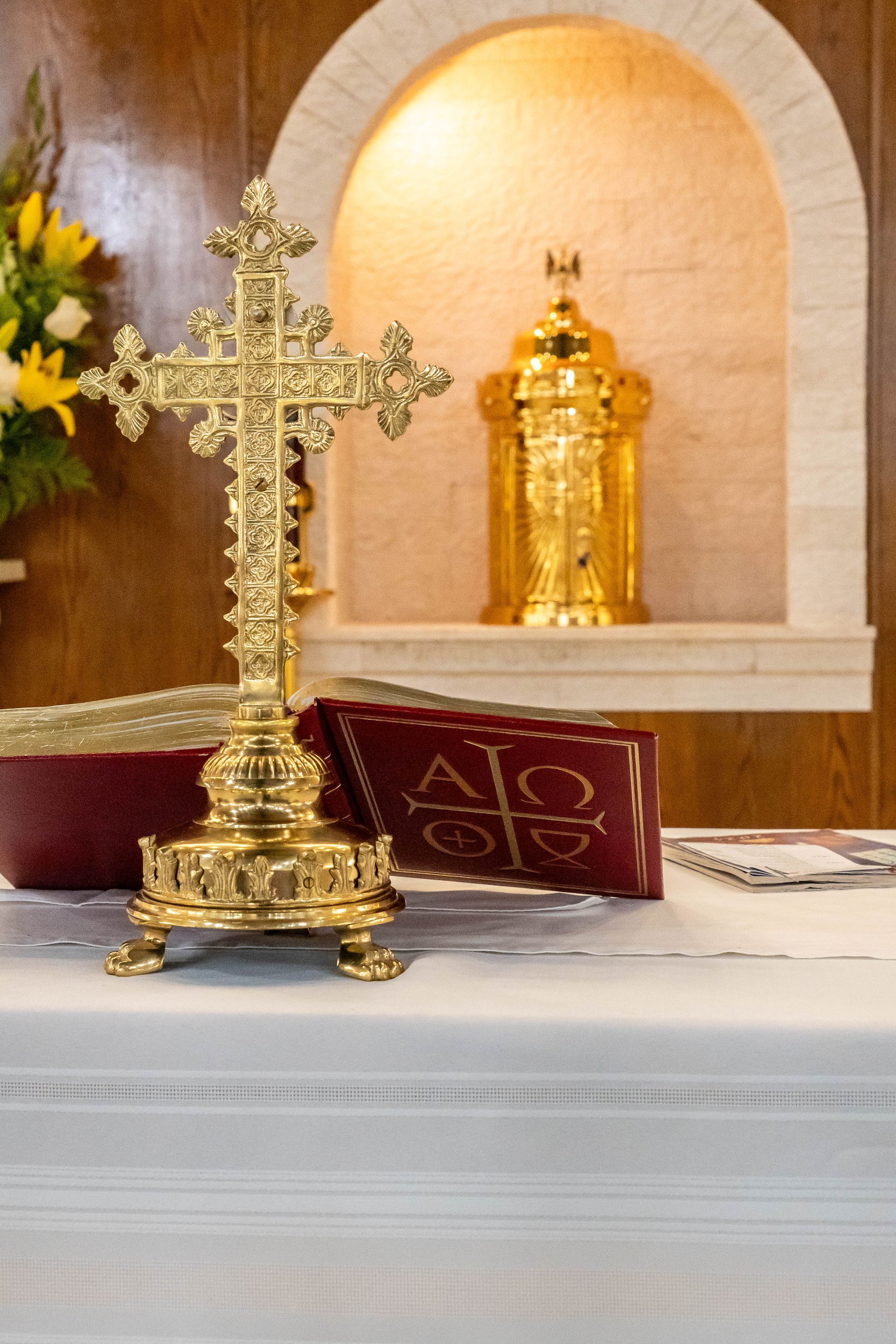 An ornate golden altar cross stands before an open red liturgical book on a white altar, with a gold tabernacle behind.