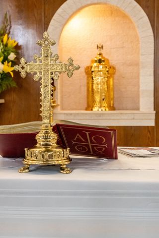 An ornate golden altar cross stands before an open red liturgical book on a white altar, with a gold tabernacle behind.