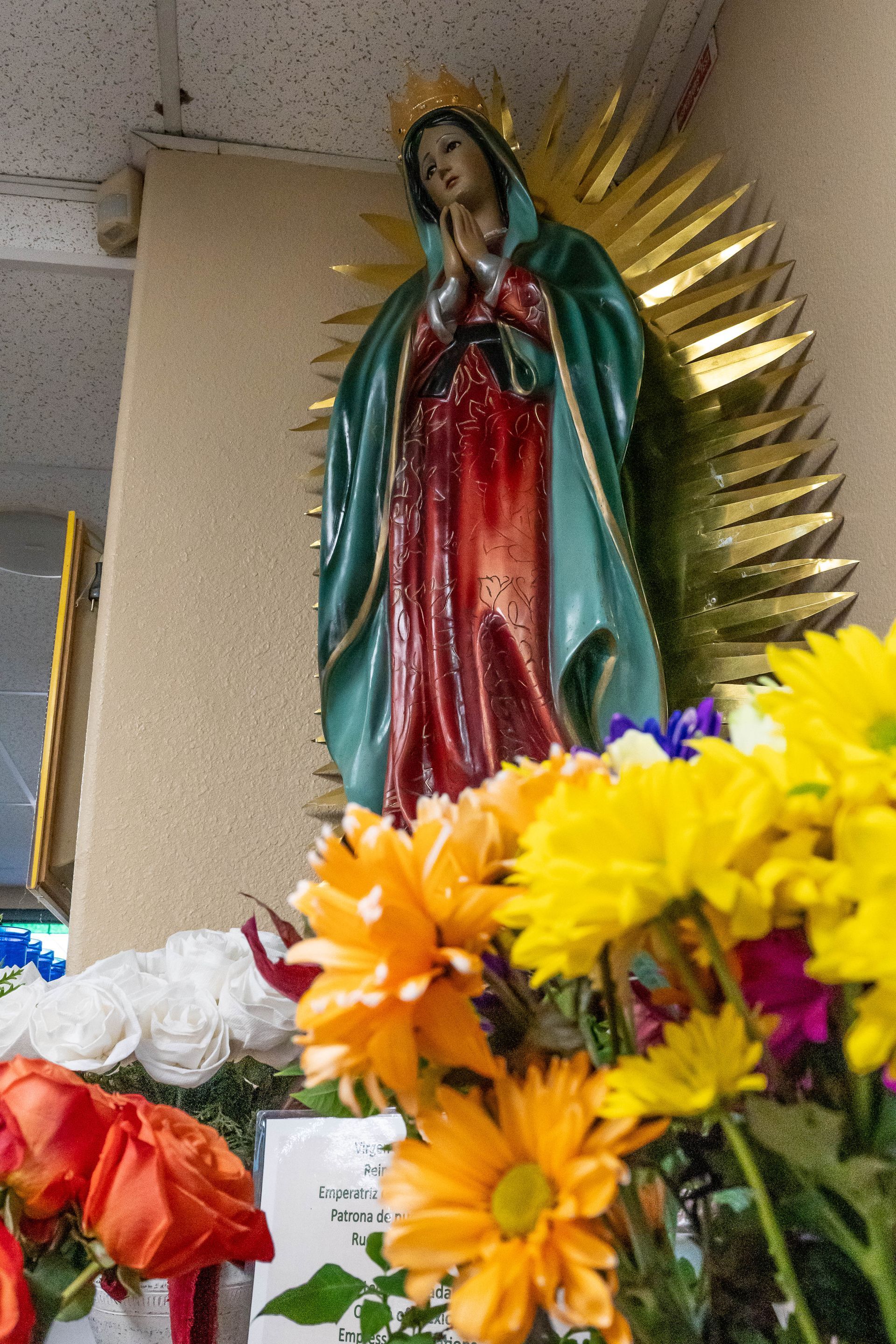A statue of Our Lady of Guadalupe set against a gold sunburst, surrounded by colorful orange and yellow flowers.