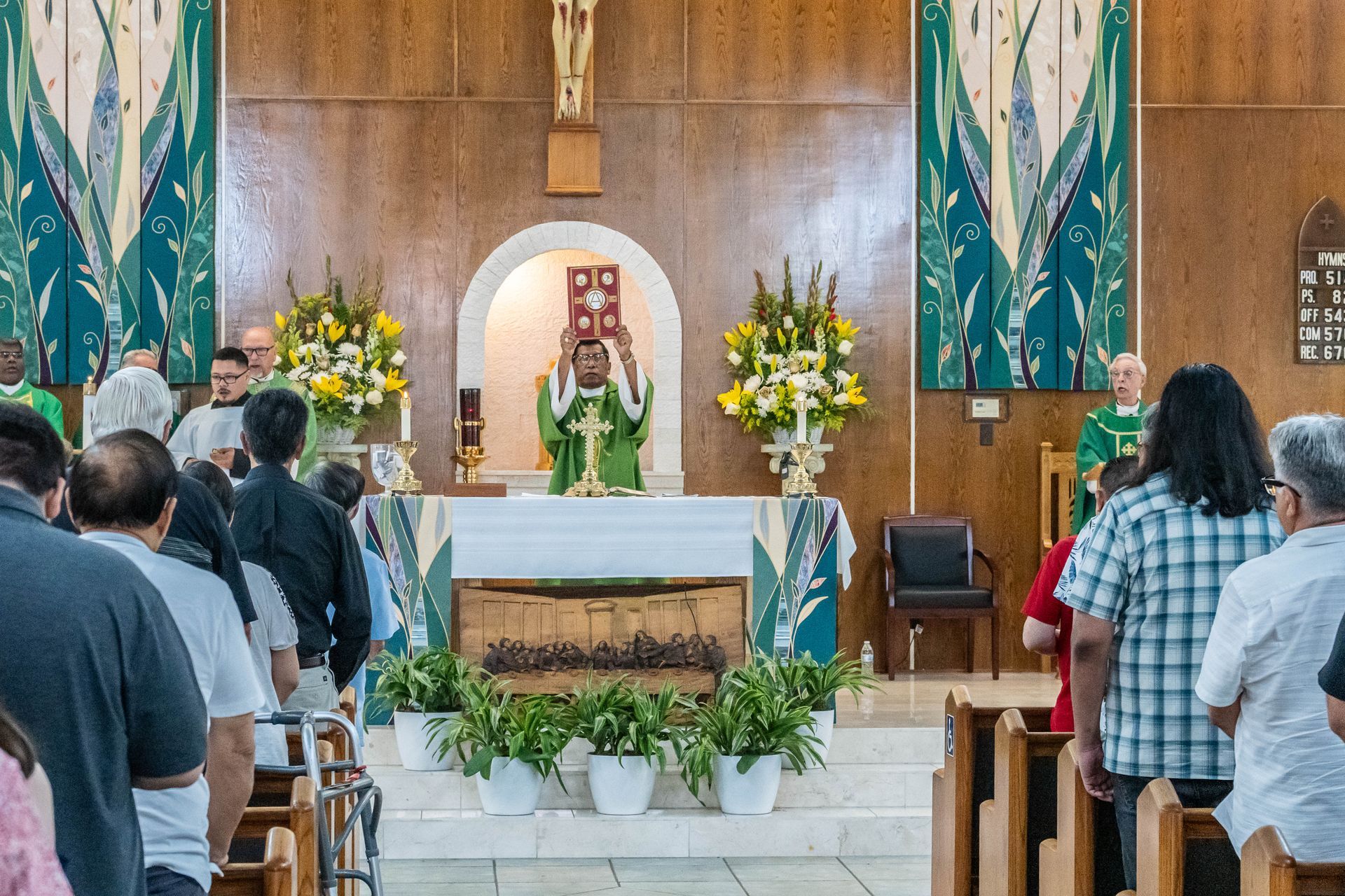 A priest in green vestments elevates a red book during a church service before a congregation.